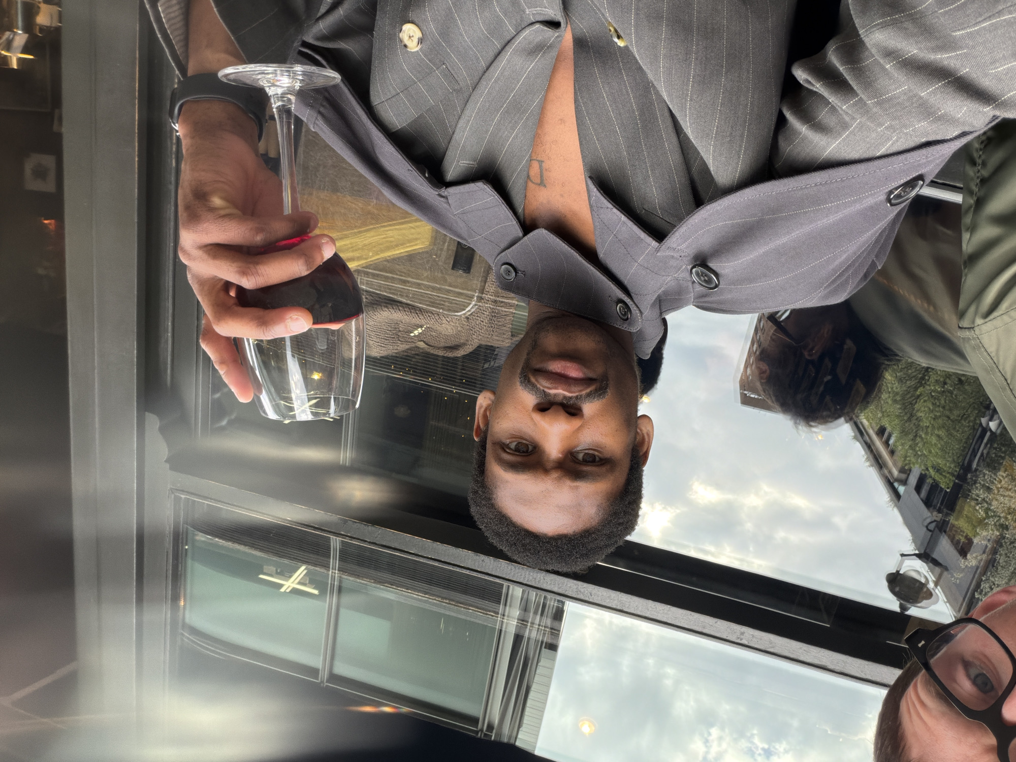 The image shows a man holding a glass of red wine, wearing a gray pinstripe suit jacket. He is indoors, sitting at a table in front of a window. Reflected in the window are buildings and a cloudy sky, suggesting a daylight setting. In the lower right corner, part of another person's face is visible, wearing glasses. The tattoo is visible on the man's chest.
The location is London, United Kingdom.