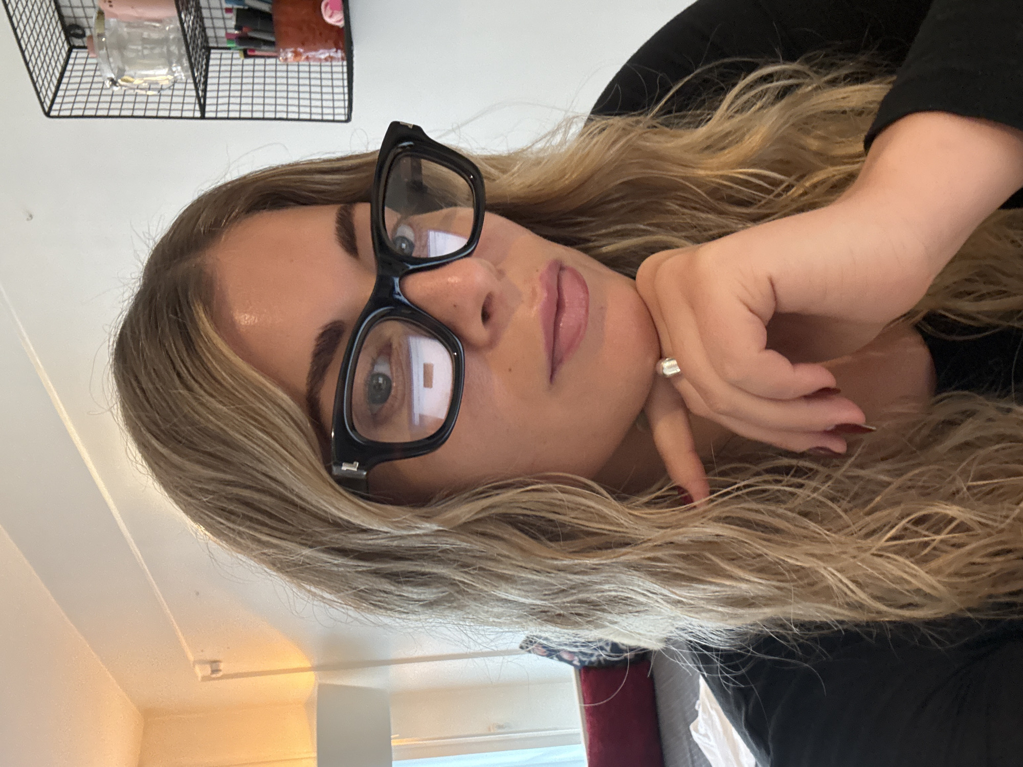 A woman with blonde hair and black-framed glasses poses indoors. Her left hand is near her face, with a ring on her finger. A black metal shelf with various items is visible in the upper left corner. The background includes a white wall and part of a window. The location within Paris, France cannot be determined from the image.