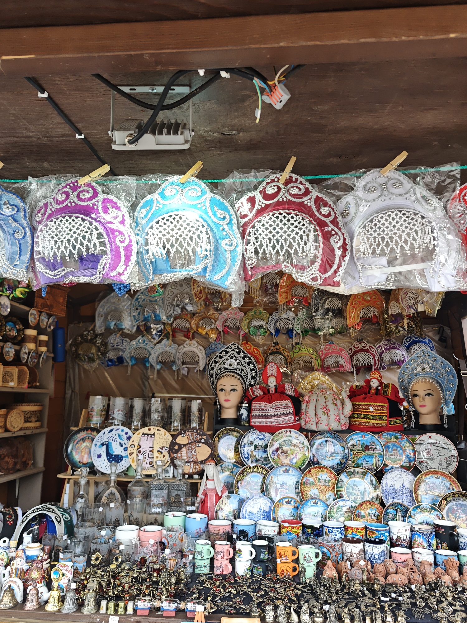 A brightly lit market stall in Uglich, Russia, displays a variety of souvenir items. At the top, a row of colorful, ornamental headdresses hangs from clothespins, above a second layer of smaller headdresses. Below this, mannequin heads wearing decorative headdresses are visible. The middle ground features a shelf of decorative plates and glassware, including mugs. The foreground is filled with small figurines and souvenirs.