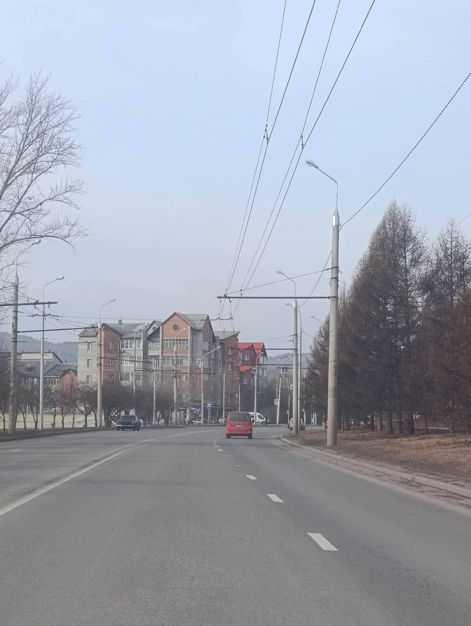 The image shows a straight section of a wide asphalt road in Krasnoyarsk, Russia. The road has two lanes in each direction, separated by white painted lines. A red minivan is traveling away from the camera in the right lane. A dark-colored sedan is approaching in the opposite direction in the left lane. Buildings line the left side of the road, a mix of gray and red brick apartments. On the right side of the road, bare trees and utility poles stretch into the distance. Overhead, a network of wires is visible. The sky is overcast, giving the scene a muted light. The image seems to be taken in early spring, with the trees lacking foliage.