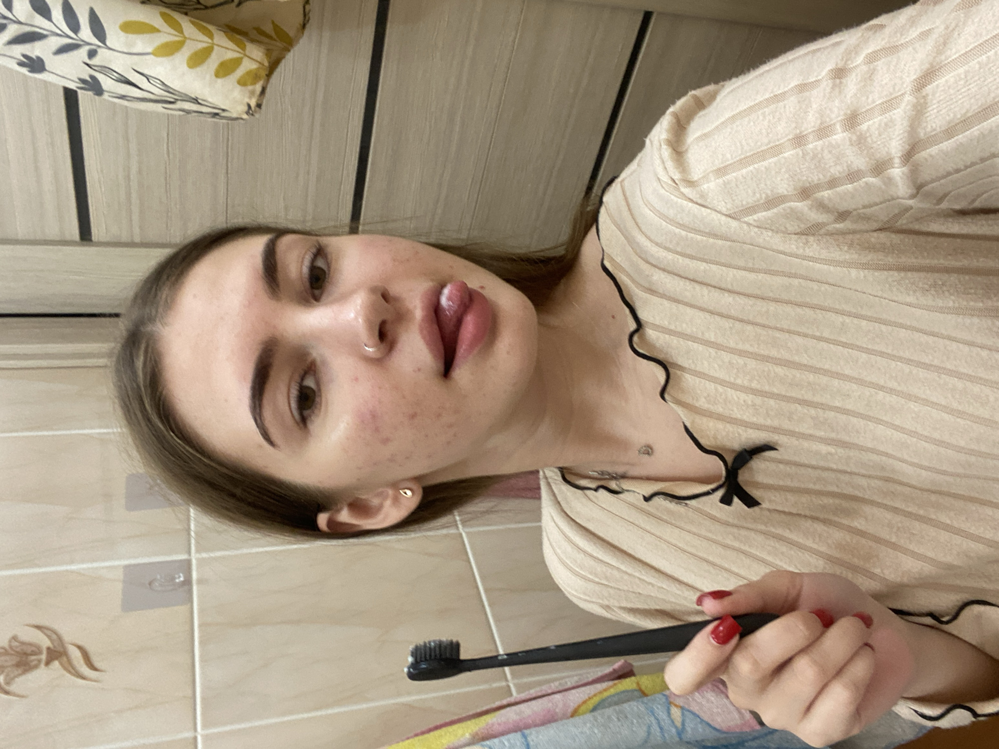 A young woman is depicted in a sideways, close-up shot, holding a black toothbrush. She is looking towards the camera with a playful expression, her tongue slightly extended. She has a nose piercing (a silver hoop) and a small gold stud earring. Her skin shows some redness and blemishes on her cheek.
She is wearing a light beige, ribbed long-sleeved top with black ruffled trim around the neckline, adorned with a small black bow on the chest. Her fingernails are painted with red nail polish.
The background consists of light-colored, possibly beige or cream, square wall tiles. One tile features a subtle floral design. Above the tiled section, there is wooden paneling, possibly a door or another wall. In the upper left corner, a patterned fabric object, possibly a pillow or blanket with dark grey and yellow leaf patterns, is visible. At the bottom right, a colorful striped towel or cloth can be seen. Two small, clear adhesive hooks are present on the tiled wall. There is also a small mark or tattoo visible on her upper chest/neck area.
