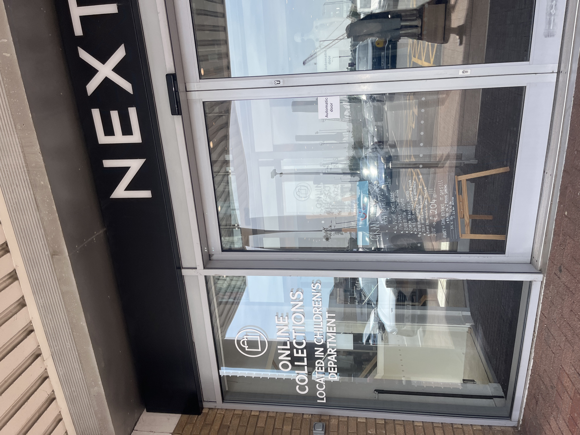 The image shows the entrance to a Next store, likely in East Suffolk, UK, based on the visible signage. The entrance has glass doors with "NEXT" branding on a black panel to the left. The lower glass panel displays "ONLINE COLLECTIONS LOCATED IN CHILDREN'S DEPARTMENT". Reflections in the glass doors indicate an outdoor environment with vehicles and other structures. A sign indicating "Automatic door" is visible on the door. A hand-written sign advertising a TUI Gran Canaria short break is taped on the door with details about dates, flights, and pricing.