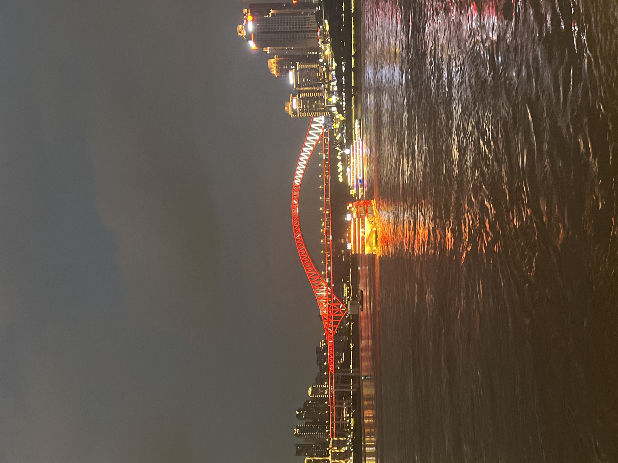 This is a nighttime photograph of the Yangtze River in Chongqing, Yuzhong District, China. A bright red, illuminated arch bridge spans the river. On the river, several boats are visible, one in the foreground with yellow lights and another with multiple colors further back. The distant cityscape is illuminated with numerous lights from buildings. The sky is dark and cloudy, with reflections of the city lights visible on the water's surface.