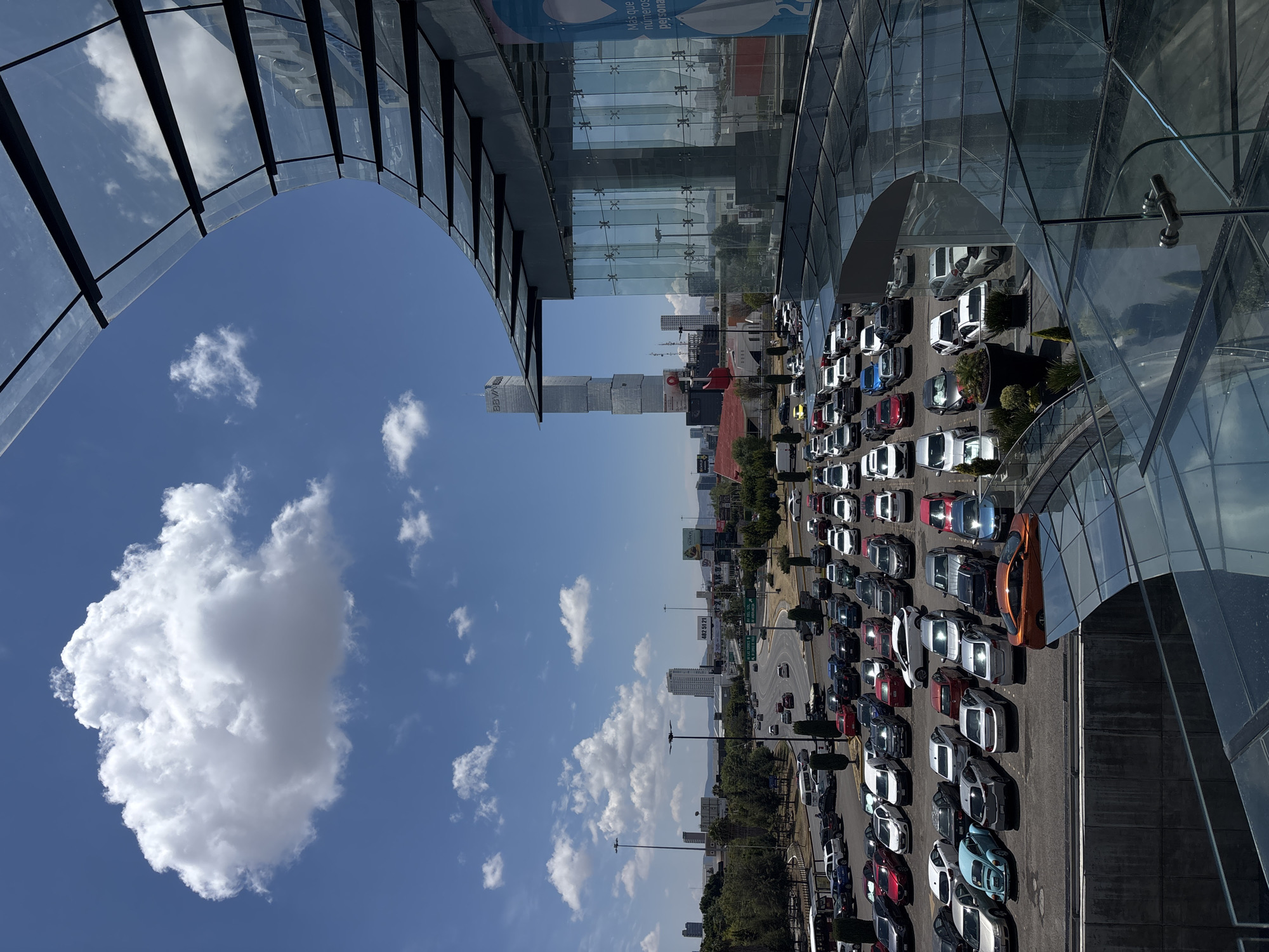 The image shows an elevated view of an urban scene in San Bernardino Tlaxcalancingo, Mexico. A modern building with a glass facade occupies the upper portion of the image. Under the building, rows of parked vehicles fill a large lot, primarily cars of various colors. Beyond the parking lot, a busy road with heavy traffic stretches into the distance, with other buildings visible along the horizon. The sky above is bright blue with scattered clouds.