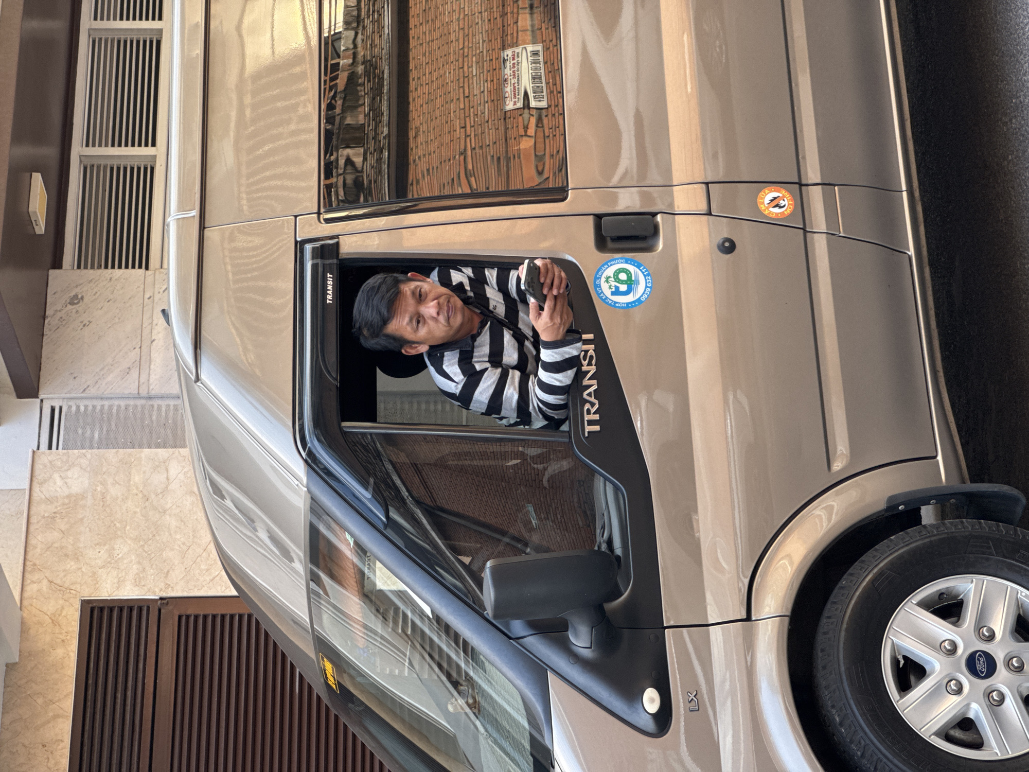 A medium shot shows a man wearing a black and white striped shirt, peering out of the driver's side window of a light brown Ford Transit van. He holds a smartphone in his right hand. The van is parked in front of a building with a marble facade and horizontal brown blinds. The background reflects buildings and trees. A blue and green sticker with Vietnamese text and a phone number is visible on the van's door. Another small circular sticker with an orange border and the words "NO FIRE" is located below the door handle. The overall lighting suggests an outdoor urban environment.