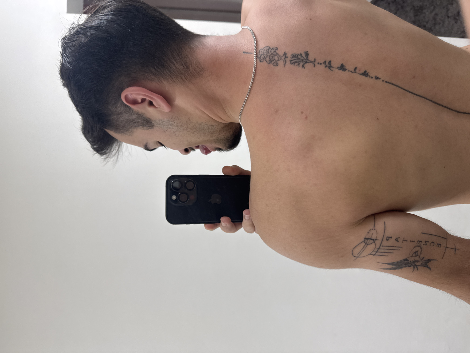 A shirtless man with dark hair and light skin is taking a selfie in front of a white wall, possibly in a bathroom. He is holding a black iPhone. He has tattoos on his back and arm. The tattoo on his back is a floral design that extends down his spine. The tattoo on his arm includes the word "PATIENCE." There is a silver chain around his neck. The location appears to be indoors. There is no indication of this image being taken in Sofia, Bulgaria.