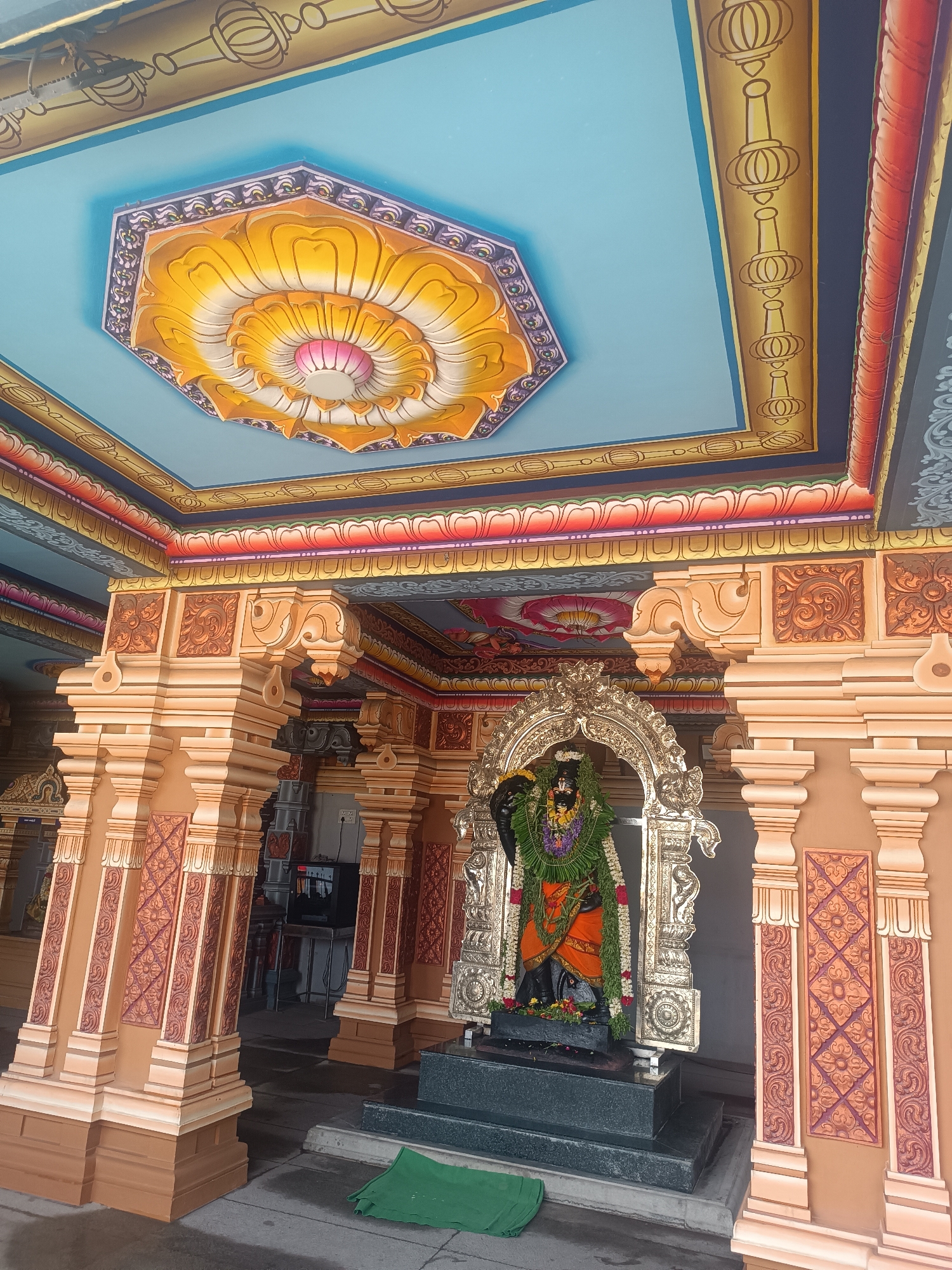 The image depicts a statue of a deity inside a Hindu temple in Ayanambakkam, India. The statue is dark-colored and adorned with garlands, and it is housed within an ornate silver or gold-colored frame. The temple's architecture features multiple pillars decorated with patterned carvings. The ceiling is painted in shades of blue, yellow, and orange, with a large lotus-shaped design at its center. There is green cloth placed in front of the statue's platform.