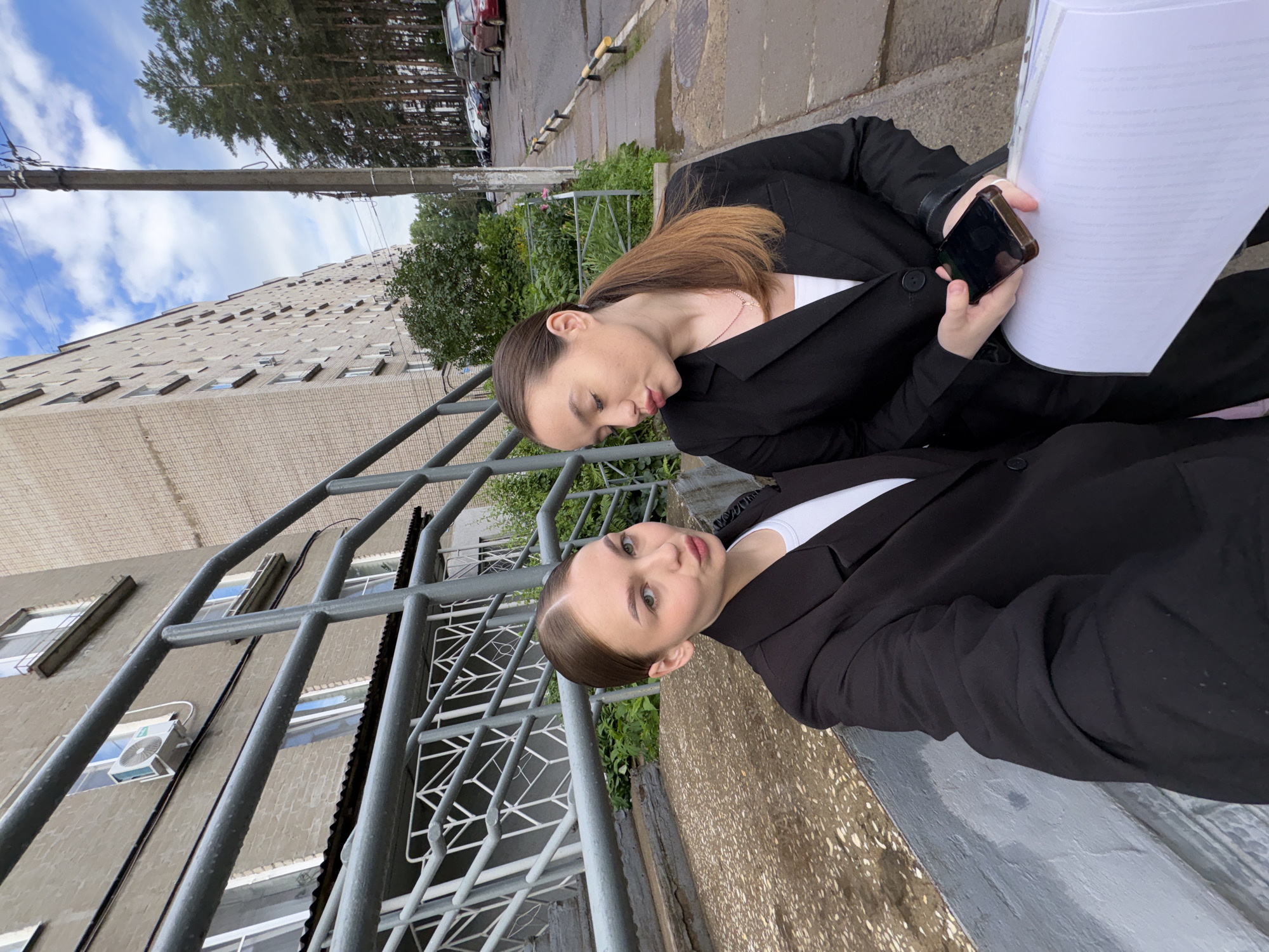 Two young women in black blazers and white t-shirts are standing on concrete stairs outdoors, with a long apartment building in the background. The woman on the left is closer to the camera and has her face turned towards it with her lips pursed. The woman on the right is looking down and to her left, holding a smartphone in her right hand and a large stack of white paper in her left. The environment appears to be a residential area in Chaikovsky, Russia, indicated by the architecture of the apartment building and the presence of parked cars and trees along the sidewalk. The sky is partly cloudy.