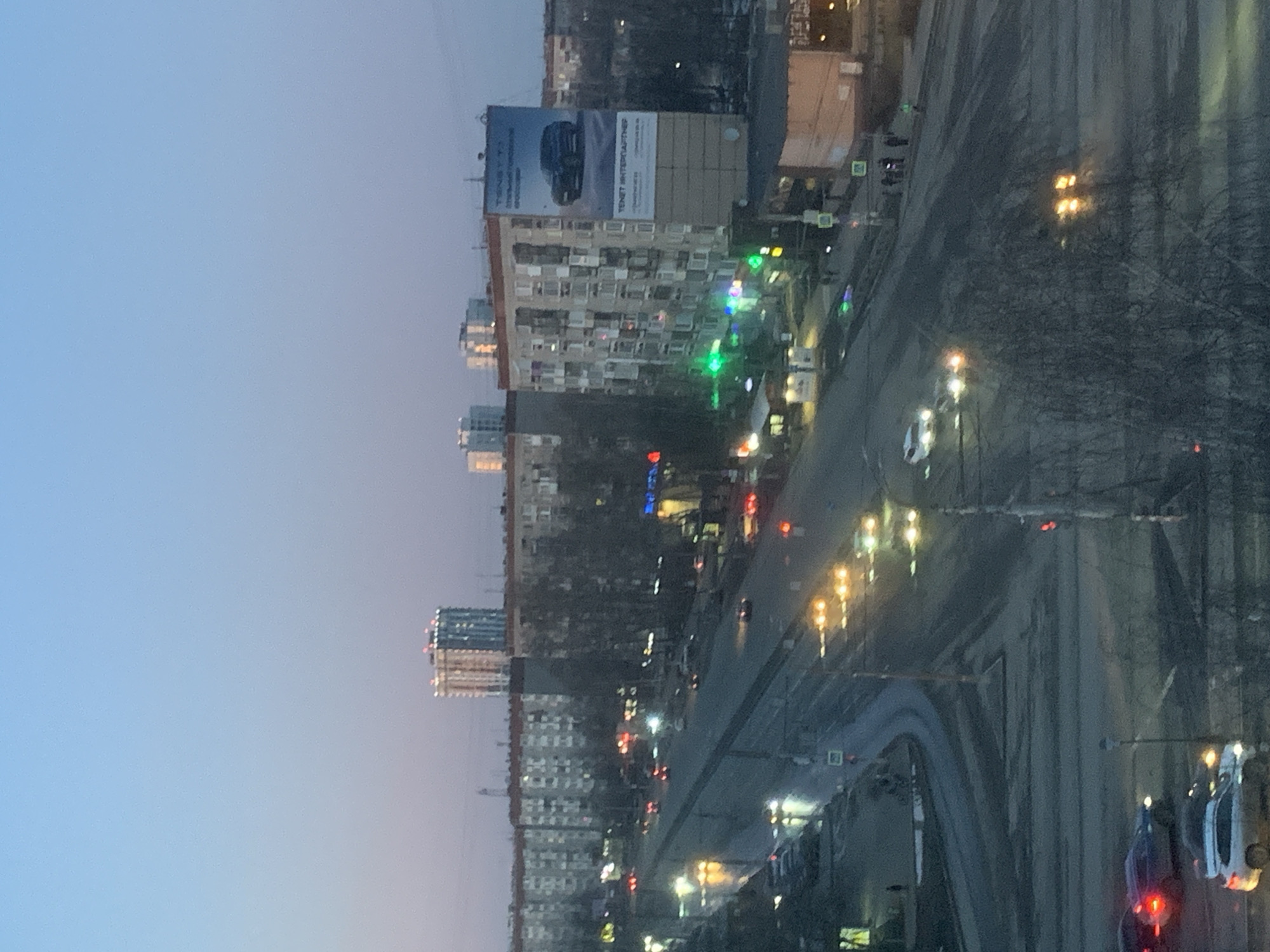 The image presents a cityscape at dusk in Izhevsk, Russia. Tall apartment buildings dominate the skyline, interspersed with trees and streetlights. A billboard tops one of the buildings. A road runs alongside the buildings, filled with cars, their headlights reflecting off the wet surface. Pedestrian crossings are visible, indicating urban traffic management. The sky transitions from a soft blue to a darker hue, suggesting the onset of evening.