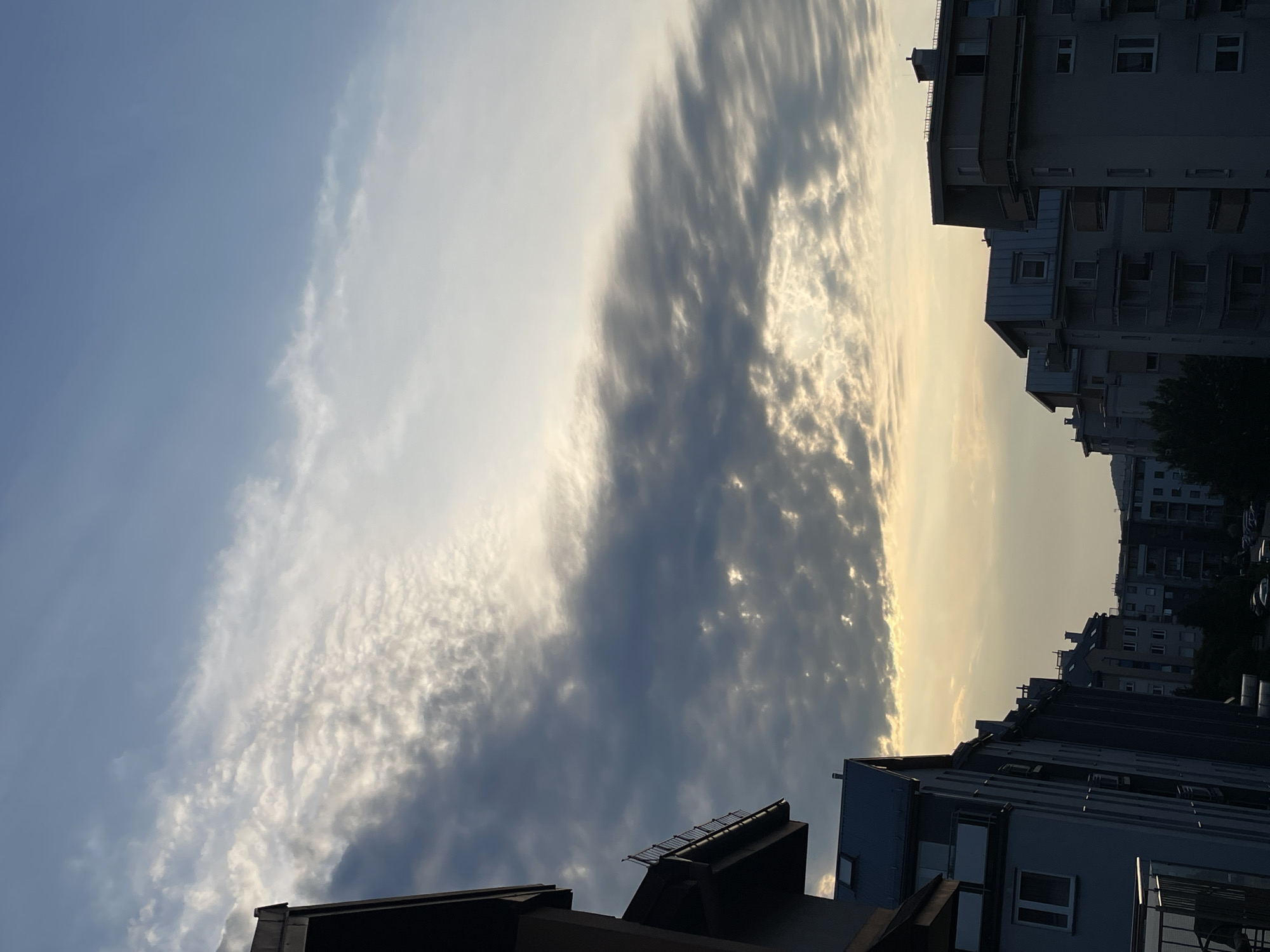 An outdoor, slightly elevated, medium shot captures a cityscape under a dramatic sky during what appears to be sunrise or sunset. In the foreground, to the left, a dark, angular building element, possibly a rooftop or balcony railing, juts into the frame. To the right and center, a series of apartment buildings with multi-story facades, balconies, and windows are visible, receding into the distance. The buildings are predominantly gray and feature a repeating architectural style. Cars are parked along the street in the lower right background. The sky is filled with a dynamic display of clouds. A prominent band of dark, textured clouds stretches horizontally across the middle of the frame, with patches of brighter, illuminated clouds interspersed. Above this, the sky transitions to a lighter blue, with wispy white clouds. The lower portion of the sky, near the horizon, is a soft yellow and orange, indicating the presence of the sun. The overall mood is one of a serene urban environment at dawn or dusk, characterized by the interplay of light and shadow on the buildings and the striking cloud formations.