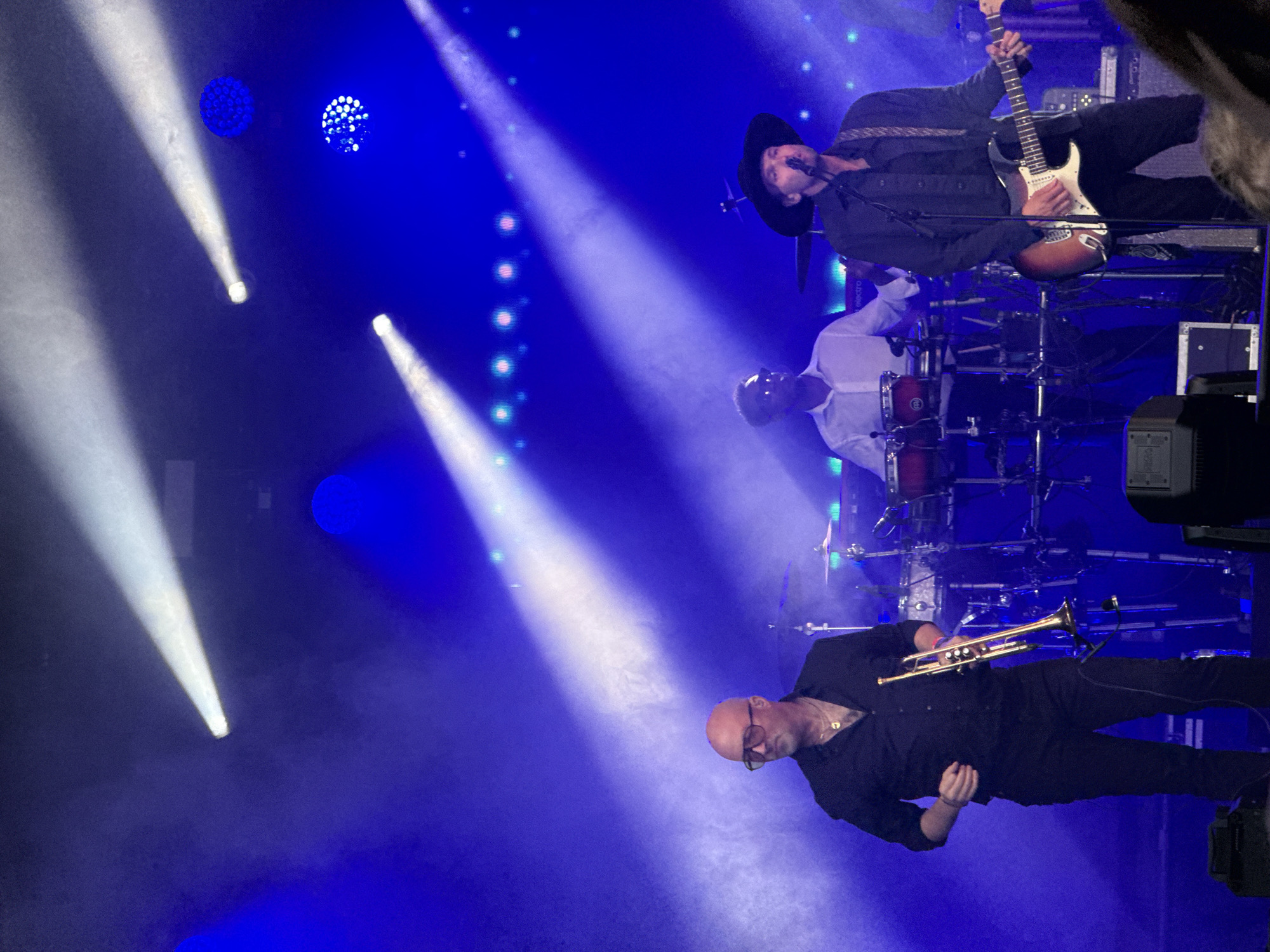 The image shows a band performing on stage in a concert venue. The stage is lit with blue and white spotlights, creating a dramatic atmosphere. In the foreground, a man wearing a black shirt and glasses is playing a trumpet. He appears focused and engaged in his performance. Behind him, a guitarist in a black hat is playing a guitar. He is also dressed in black and appears to be singing or speaking into a microphone. A drummer is visible behind the guitarist, with drums and cymbals. The overall scene suggests an energetic live music event. The colors are vibrant, with the blue stage lighting dominating the scene. The audience is not visible, but the presence of a full band and stage lighting indicates a public performance. There is no visible text in the image.
