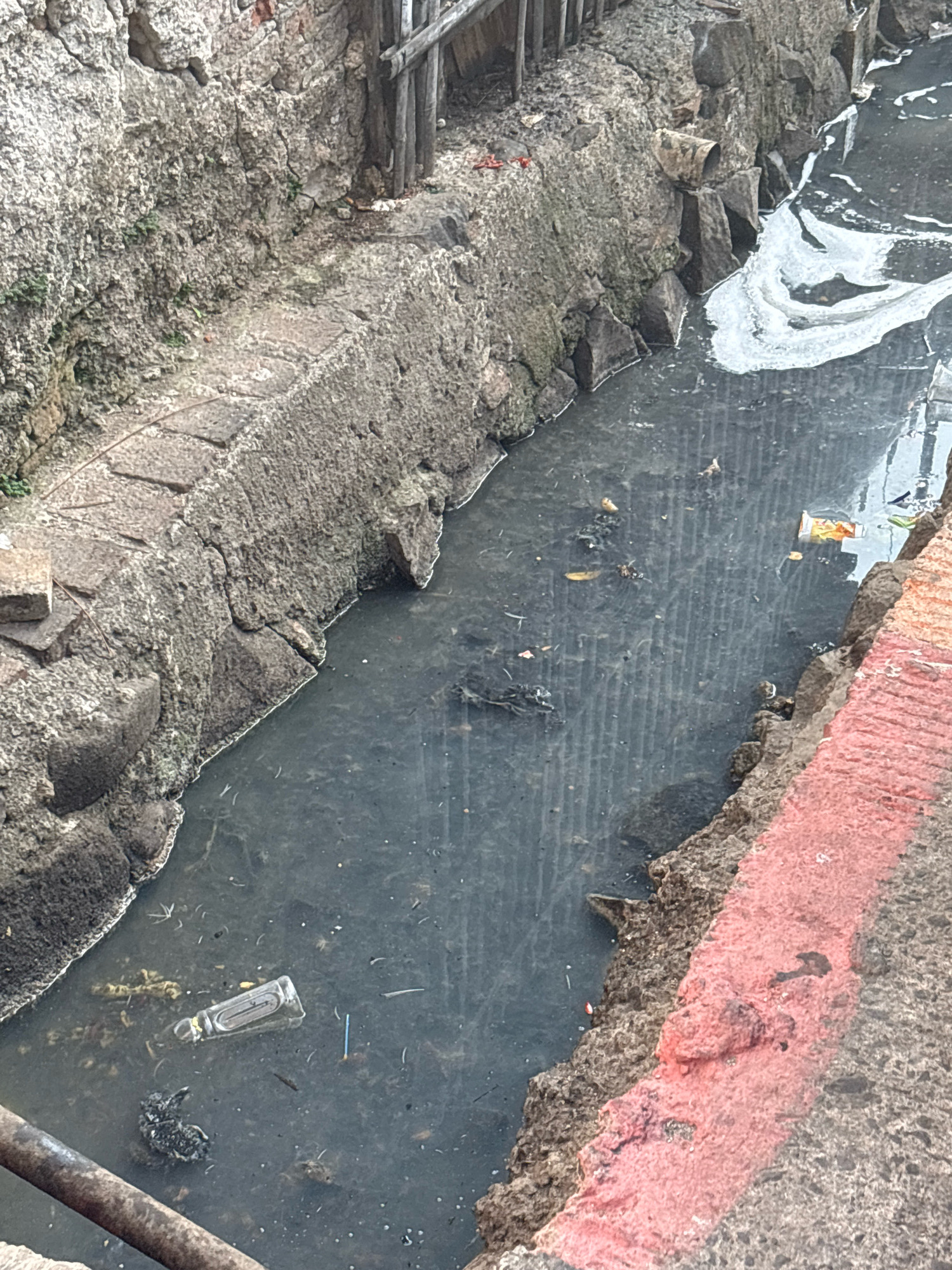 The image depicts a polluted waterway, likely a drain or canal, within the city of North Jakarta, Indonesia. The water is dark and murky, with visible debris and foam on the surface. The banks of the waterway are made of rough, textured stone and concrete, some sections of which are crumbling. There are pieces of trash scattered in and around the water, including a plastic bottle, a carton, and other unidentifiable objects. A red line is painted on the edge of the concrete structure on the right side of the image. The overall environment suggests neglect and poor sanitation.