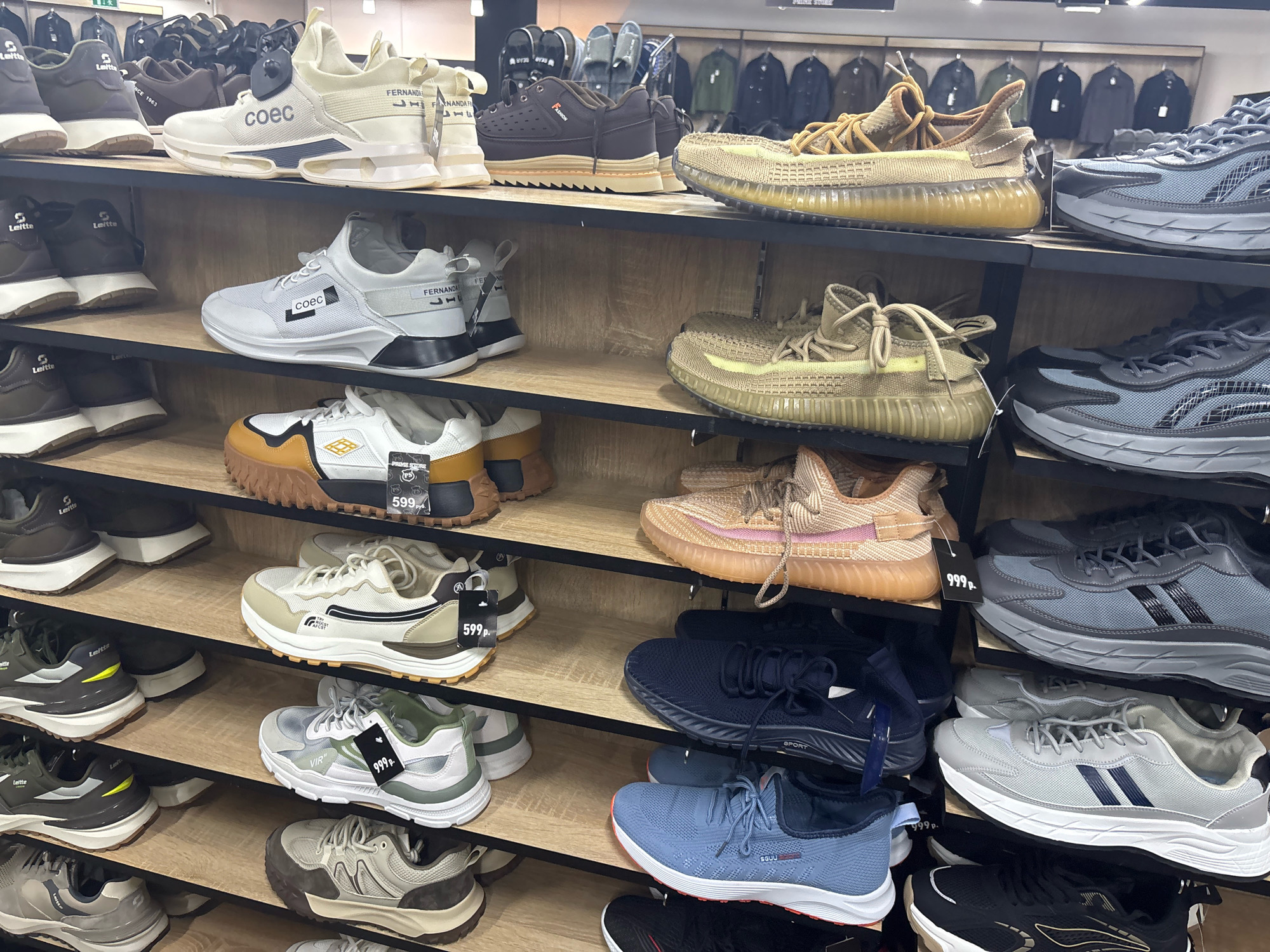 The image shows a retail display of athletic footwear in a store. The shoes are arranged on wooden shelves. The brands and models are varied, and several pairs have price tags. The background shows more clothing racks with jackets and possibly other apparel. The overall environment appears to be a well-lit shoe store. This specific display of athletic footwear is located at the "Sportmaster" store in the "XL" shopping center in Mytishchi, Russia.