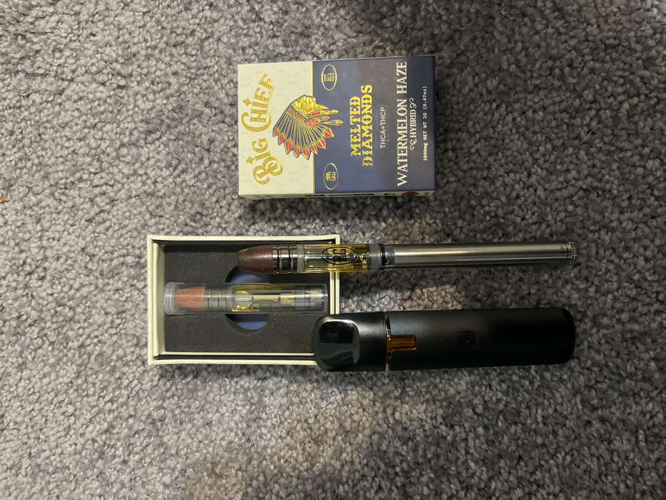 The scene is located in the city of Fox Lake, United States of America. The image displays four objects arranged on a grey, short-pile carpet. On the right, a rectangular cardboard box is visible, featuring "Big Chief" branding, an illustration of a Native American headdress, and text indicating "MELTED DIAMONDS THCA+THCP WATERMELON HAZE HYBRID 2000MG NET WT 2G (0.07oz)". To the left, an open, off-white rectangular box contains one clear cylindrical vape cartridge with amber-colored liquid and an orange-brown tip, secured in a dark interior insert. Below this open box and slightly to the right, a silver cylindrical vape pen is present, with an attached clear cylindrical vape cartridge containing amber-colored liquid and a dark brown tip, marked "CC". To the left of the silver vape pen, a black, sleek, elongated oval-shaped vaping device with a transparent amber-colored reservoir at its upper section is positioned. No individuals are present in the scene.