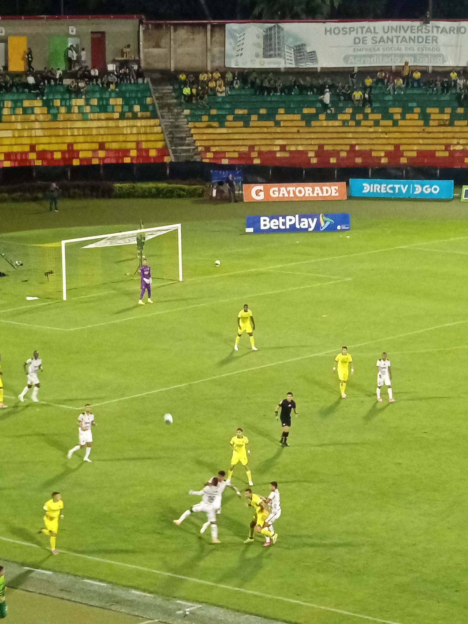 The image depicts a soccer match taking place at night in an outdoor stadium. The field is green grass, marked with white lines. Several players are visible on the field, some in white uniforms and others in yellow uniforms. One player in a purple uniform appears to be a goalkeeper, standing near a goalpost. Spectators are seated in the stands, which are sectioned with red, yellow, and green seating. Advertising banners are visible above the stands, including one for "HOSPITAL UNIVERSITARIO DE SANTANDER" and others for "GATORADE," "DIRECTV," and "BetPlay." The scene is illuminated by stadium lights, creating shadows on the field. The stadium is located in Bucaramanga, Colombia.