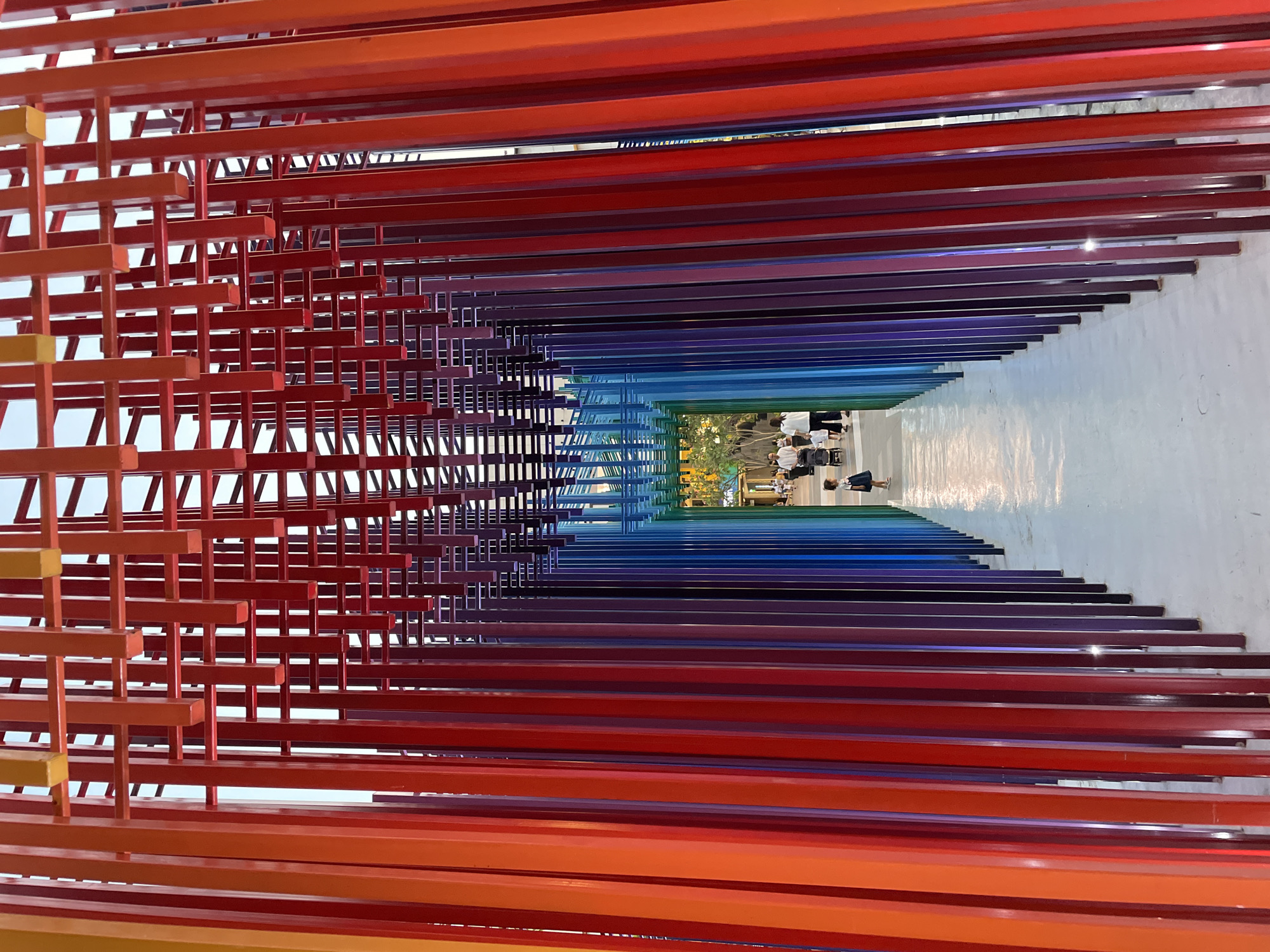 From a low vantage point in North Jakarta, Indonesia, the photograph captures a passage defined by densely arranged, parallel, rainbow-colored beams. At the entrance, on the left, these beams form an open grid structure, painted in shades of orange and red. This structure transitions to individual, closely spaced beams in a spectrum of red, purple, blue, and green. The beams create a tunnel-like effect leading to a brightly lit open space at the far end. Several people are visible in this open area, some standing and others walking. The floor beneath the beams is white, and the overall impression is one of an art installation or architectural feature.