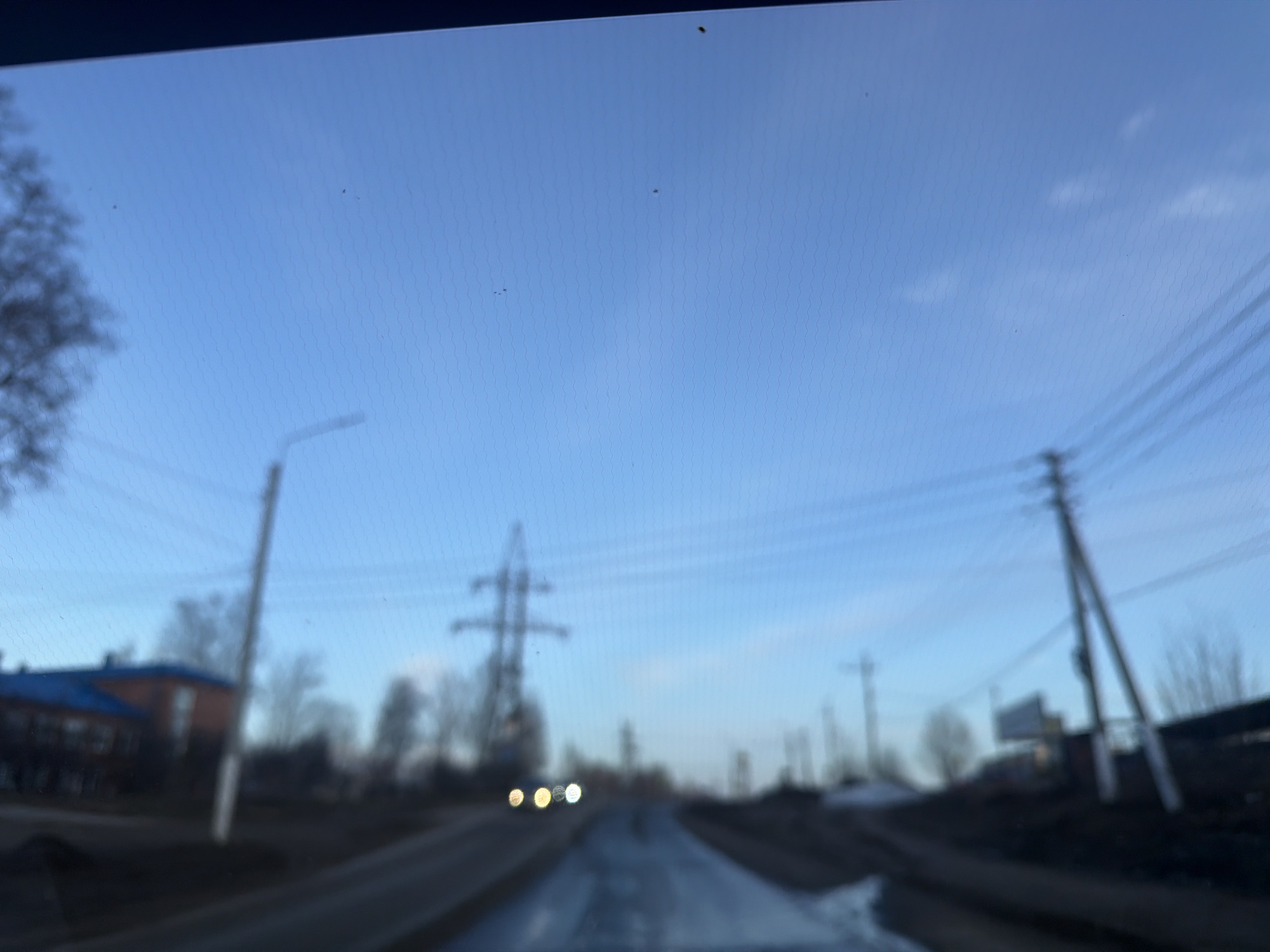 The image shows a blurry view of a road in a town, possibly the city of Chaikovsky, Russia, under a blue sky with scattered clouds. An electric power tower is visible in the distance, flanked by lamp posts and telephone poles lining either side of the road. On the left, there appears to be a building with a blue roof. The road surface is wet, reflecting light. Approaching headlights suggest a vehicle moving toward the camera, in the same direction.