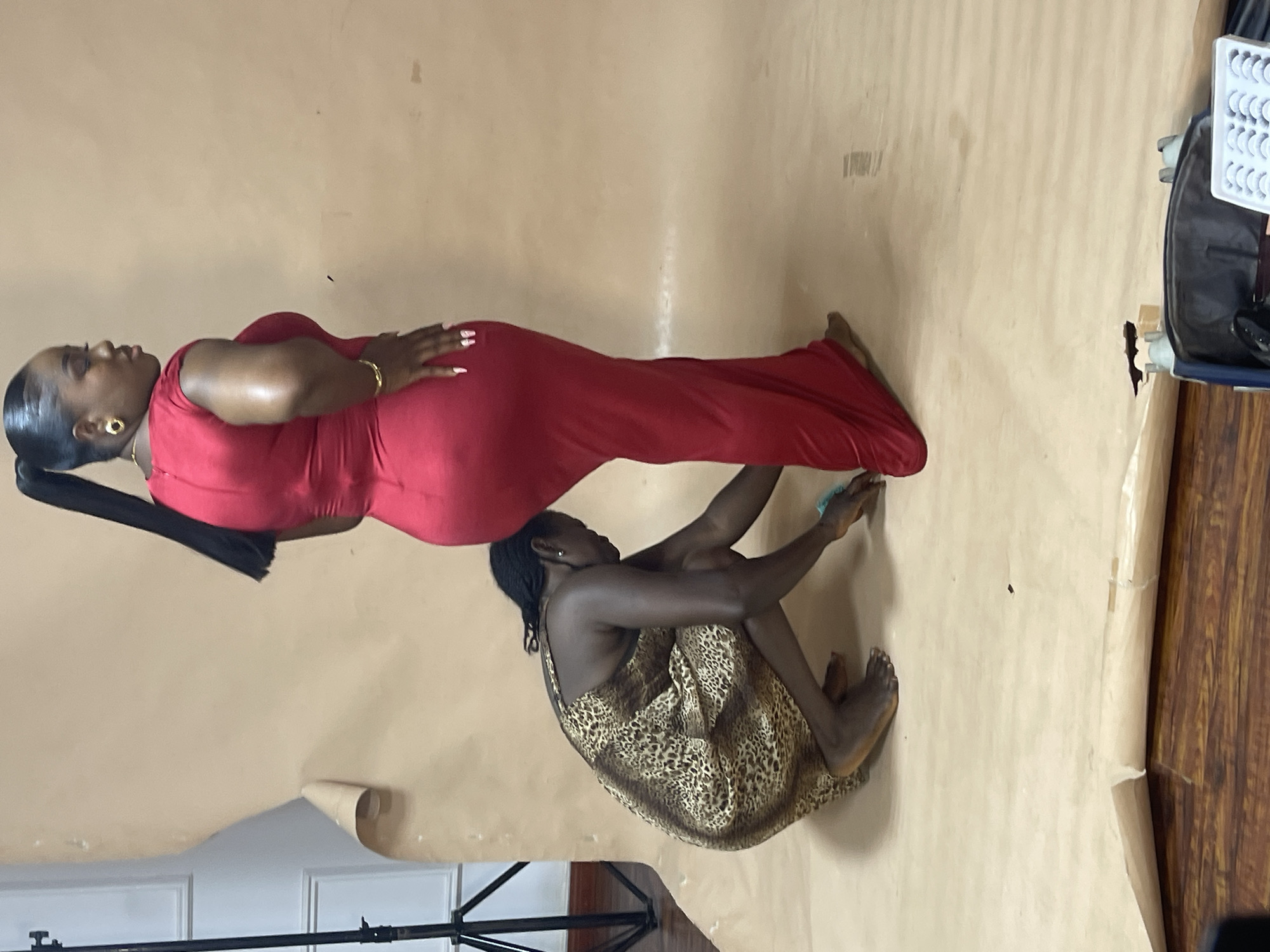 The scene depicts two dark-skinned women on a brown backdrop. One woman is lying prone, wearing a red, form-fitting dress. She has a long, black ponytail, gold earrings, and is wearing a gold bracelet. The other woman is in a seated, curled-up position, wearing an animal-print dress. Part of a light stand and a wooden-like floor are visible at the bottom. A small white eyelash case is in the right top corner. The location is most likely a studio setting in Abuja, Nigeria.