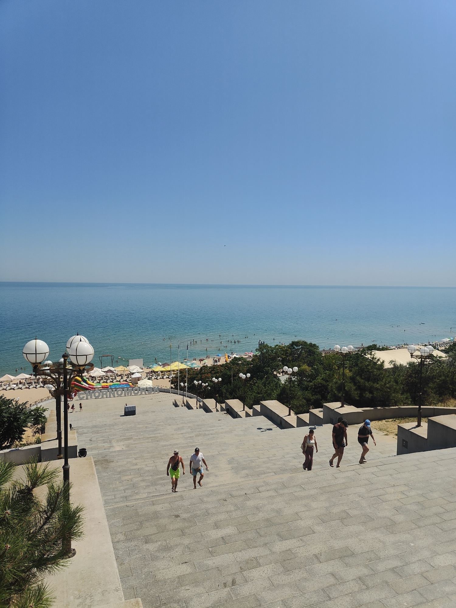 The image captures a view of the Black Sea coastline in Chornomorsk, Ukraine, from a raised vantage point. In the foreground, a wide, gray stone staircase descends towards a beach area. Several people are ascending or descending the stairs. On the staircase, two men in casual attire walk upwards, followed by a woman in a white top and patterned pants, a man in a dark shirt and shorts, and another man in a dark shirt and blue cap.
Below the staircase, a sandy beach is visible, lined with numerous beach umbrellas and lounge chairs. The sea is calm and a deep blue, with many people swimming and sunbathing. In the middle ground, large white letters spell out "CHORNOMORSK." Inflatable attractions, including a slide, are set up on the sand near the water. Lush green trees and vegetation flank the beach and rise behind it. The sky is a clear, bright blue. Two streetlights with multiple globes are positioned on the left side of the staircase, and another similar streetlight is visible further down. The overall scene depicts a sunny day at a popular coastal resort.
