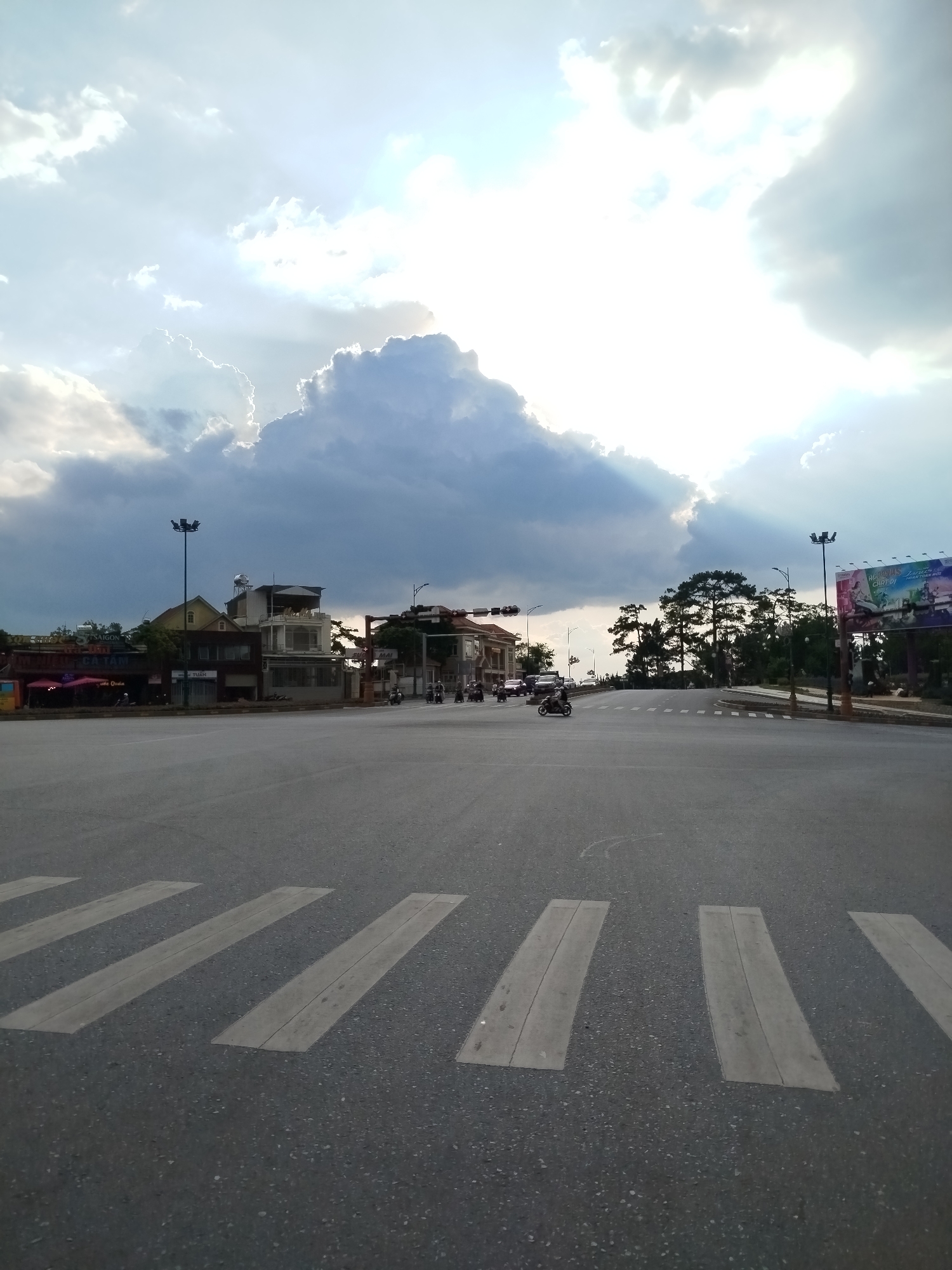The image depicts an intersection in Da Lat, Vietnam, under a cloudy sky. The sun is visible through the clouds, creating a bright, almost blinding light. The road is grey asphalt with white pedestrian crossing stripes. Several motorcycles and cars are driving on the road, moving in different directions. Buildings are visible on either side of the road, including one with a red awning and another that is multi-story with a yellow roof. There are lampposts on both sides of the road and a large billboard on the right. Trees are also visible on the right.