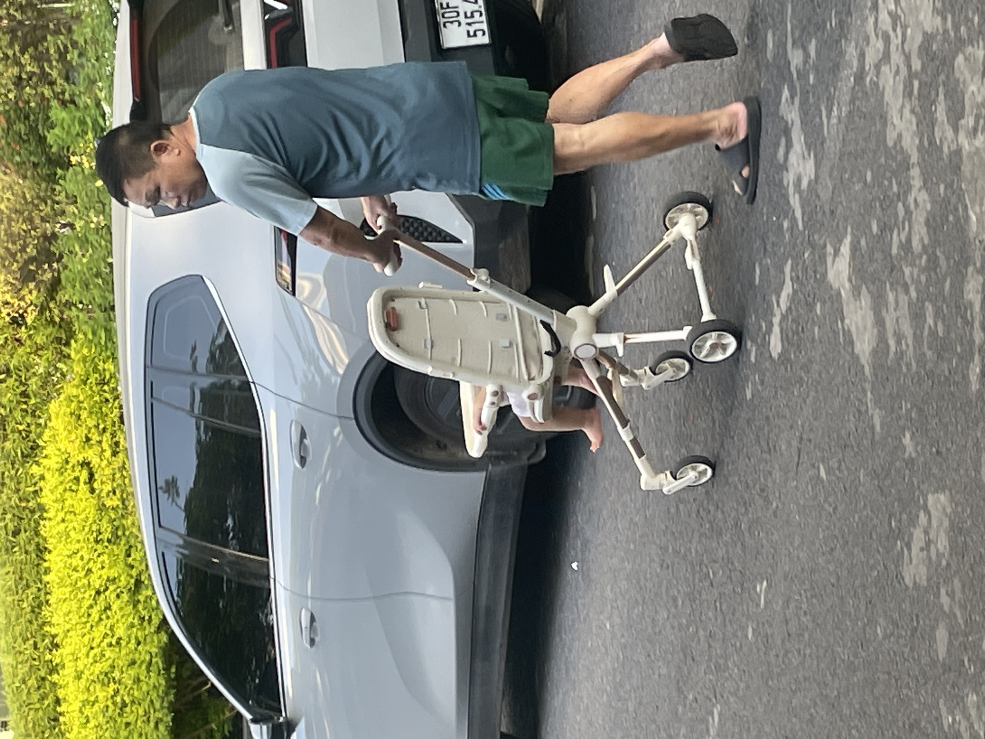 A man is pushing a stroller with a child in it down a paved road. The man is wearing a teal shirt and green shorts. The child is wearing a white outfit and is barefoot. A silver car is parked on the side of the road behind the stroller. There is a lush green hedge in the background. The license plate of the car reads "30F 515.4". The image appears to have been taken in Bac Nha Trang Ward, Vietnam.