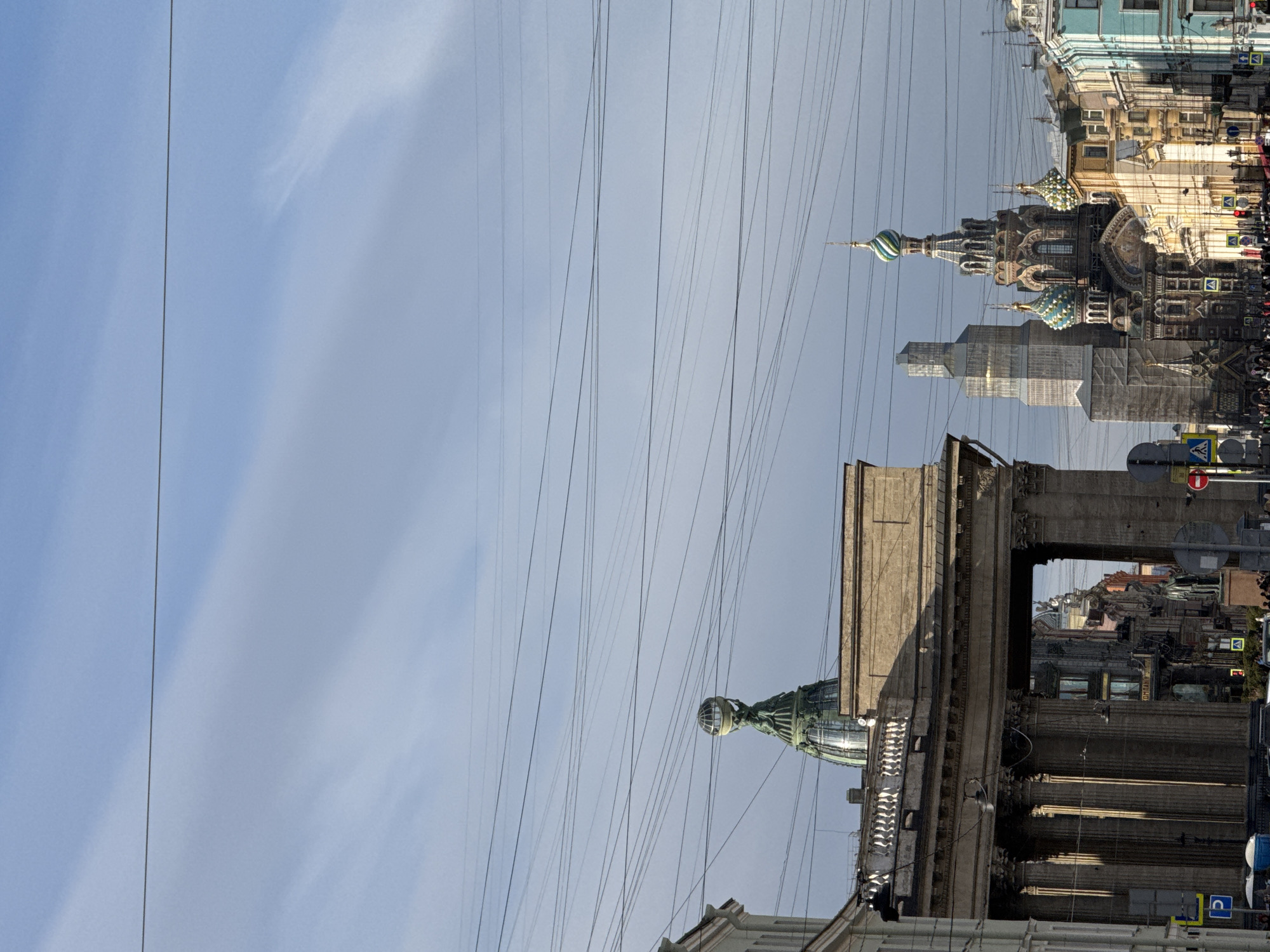 The image shows a view of Saint Petersburg, Russia, with overhead power lines dominating the sky. In the background, the Church of the Savior on Spilled Blood is visible, partly obscured by scaffolding. In the foreground, architectural details of the Singer House (also known as the House of Books) are seen, with a glimpse of its distinctive dome. The perspective is slightly tilted, enhancing the angle of the power lines against the buildings. Street signs are present, along with other buildings lining the street. People appear as indistinct figures in the background. The sky is light blue with some clouds.