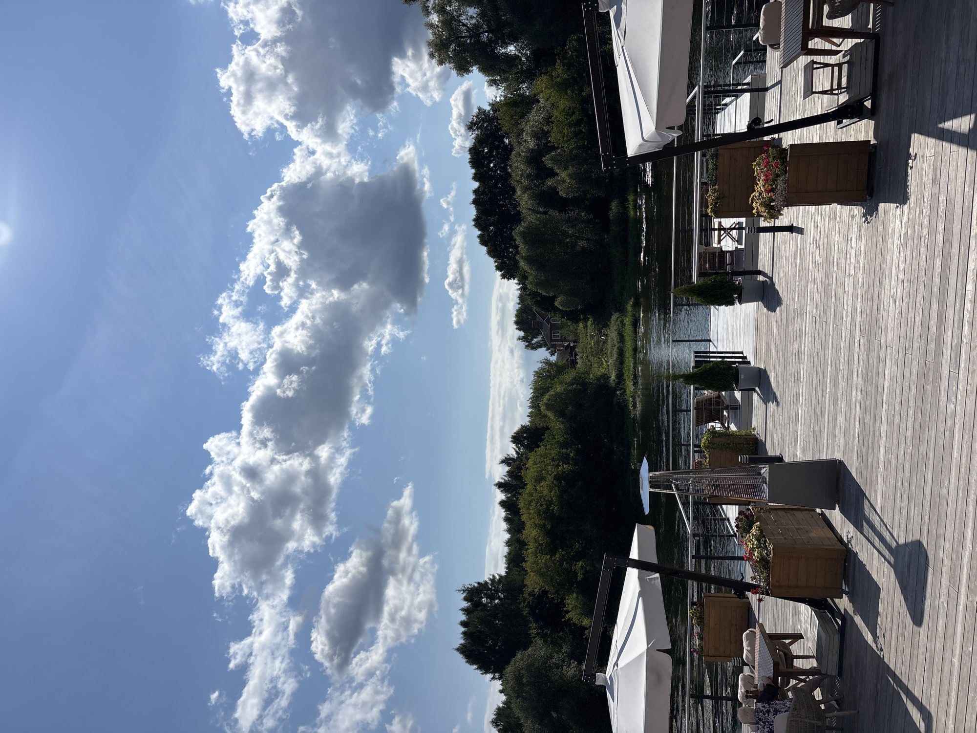 The image depicts an outdoor seating area at a waterfront establishment, likely a restaurant or cafe, in Zvenigorod, Russia. The scene is set on a wooden deck overlooking a tranquil river bordered by lush green trees. White umbrellas provide shade for tables and chairs arranged on the deck. Several wooden planters filled with flowers and small evergreen trees are strategically placed throughout the area. A tall patio heater stands near one of the seating areas. In the background, the river flows gently, reflecting the blue sky dotted with fluffy white clouds. The overall atmosphere is serene and inviting, suggesting a pleasant day for outdoor dining.
