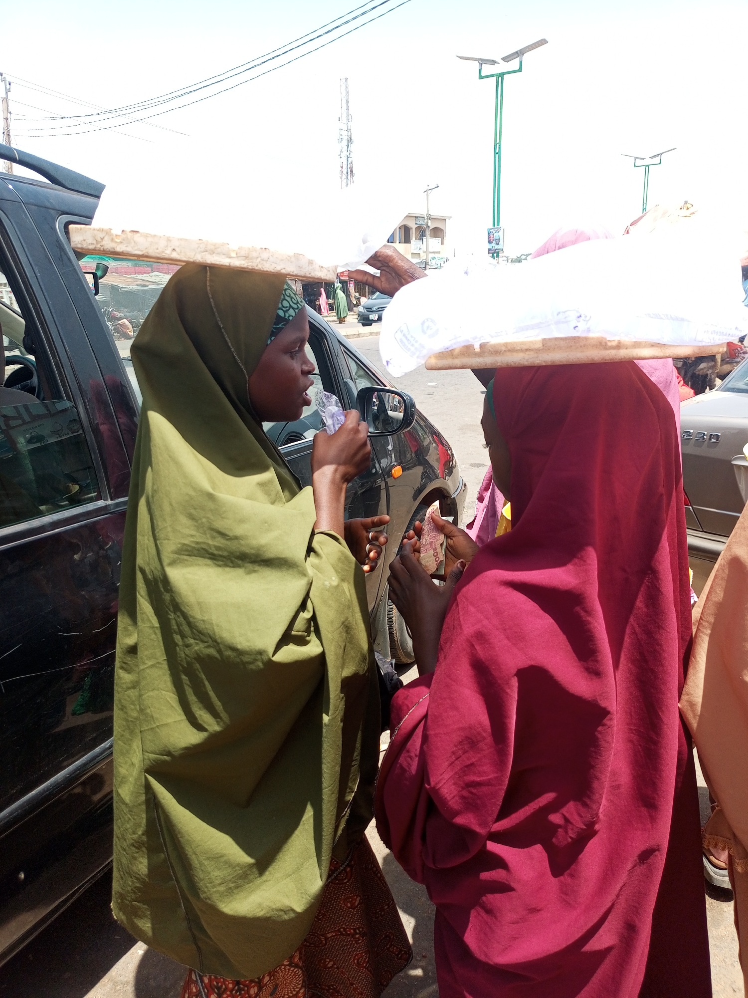 In Gombe, Nigeria, two young women wearing hijabs stand by a car, appearing to be engaged in a transaction. The woman on the left, dressed in a green hijab, holds a clear plastic bag with water and is drinking from it. The woman on the right, in a maroon hijab, holds out what appears to be money. Both women have wooden boards balanced on their heads, supporting large, white, tarp-wrapped bundles. The background suggests an urban environment with buildings, streetlights, and other vehicles. The lighting indicates it is daytime with clear skies.