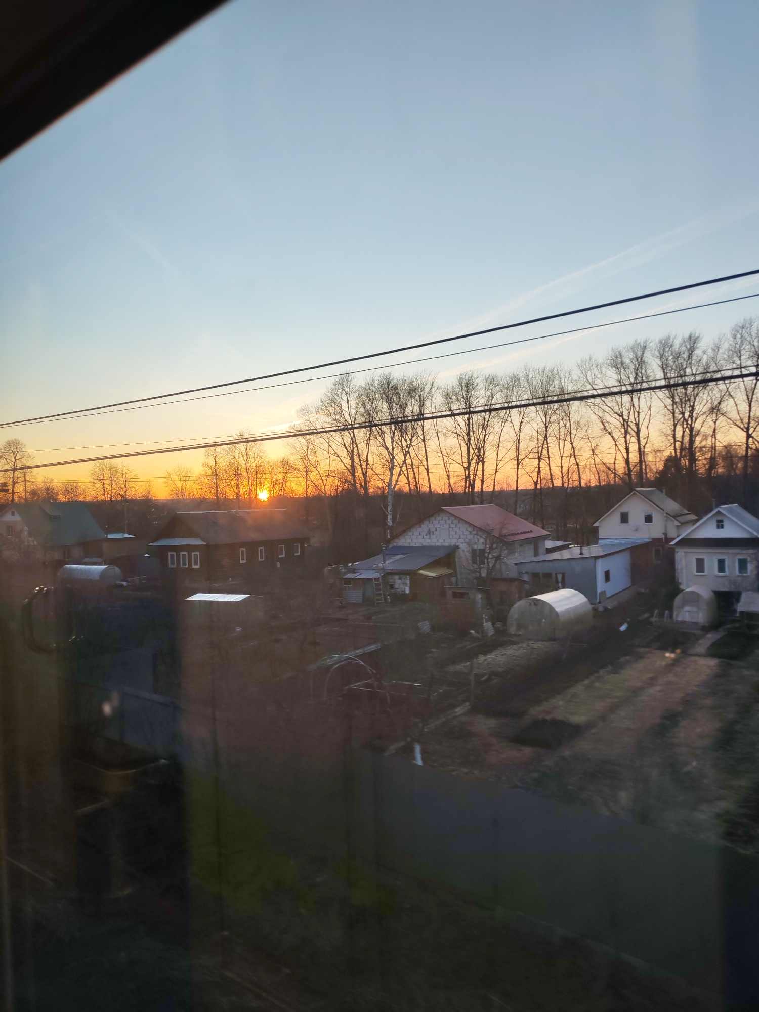 The image captures a rural or semi-rural landscape at either sunrise or sunset, viewed through a window, possibly from a train or car. The sky is dominated by a warm orange glow along the horizon, where the sun is low, silhouetting a dense line of bare deciduous trees. Above the orange, the sky transitions to a clear, pale blue, with faint white streaks, possibly contrails or wispy clouds.
In the mid-ground, a collection of houses with various architectural styles and roof colors (red, gray, light blue) are visible. One prominent dark-colored house near the center-left features three white rectangular markings vertically aligned on one section and three horizontally aligned, forming "000", on another. Several small, translucent, arch-shaped structures, likely greenhouses or polytunnels, are scattered among cultivated plots of land around the houses. Two white, cylindrical tanks are visible near one of the houses on the left.
Multiple dark power lines stretch horizontally across the upper portion of the frame, partially obscuring the sky and distant trees. The foreground and parts of the scene are affected by reflections on the window, showing dark, indistinct shapes that suggest the interior of the viewing vehicle, such as a window frame on the left. No people are visible. The overall atmosphere is calm, characterized by the soft, directional light of dawn or dusk.