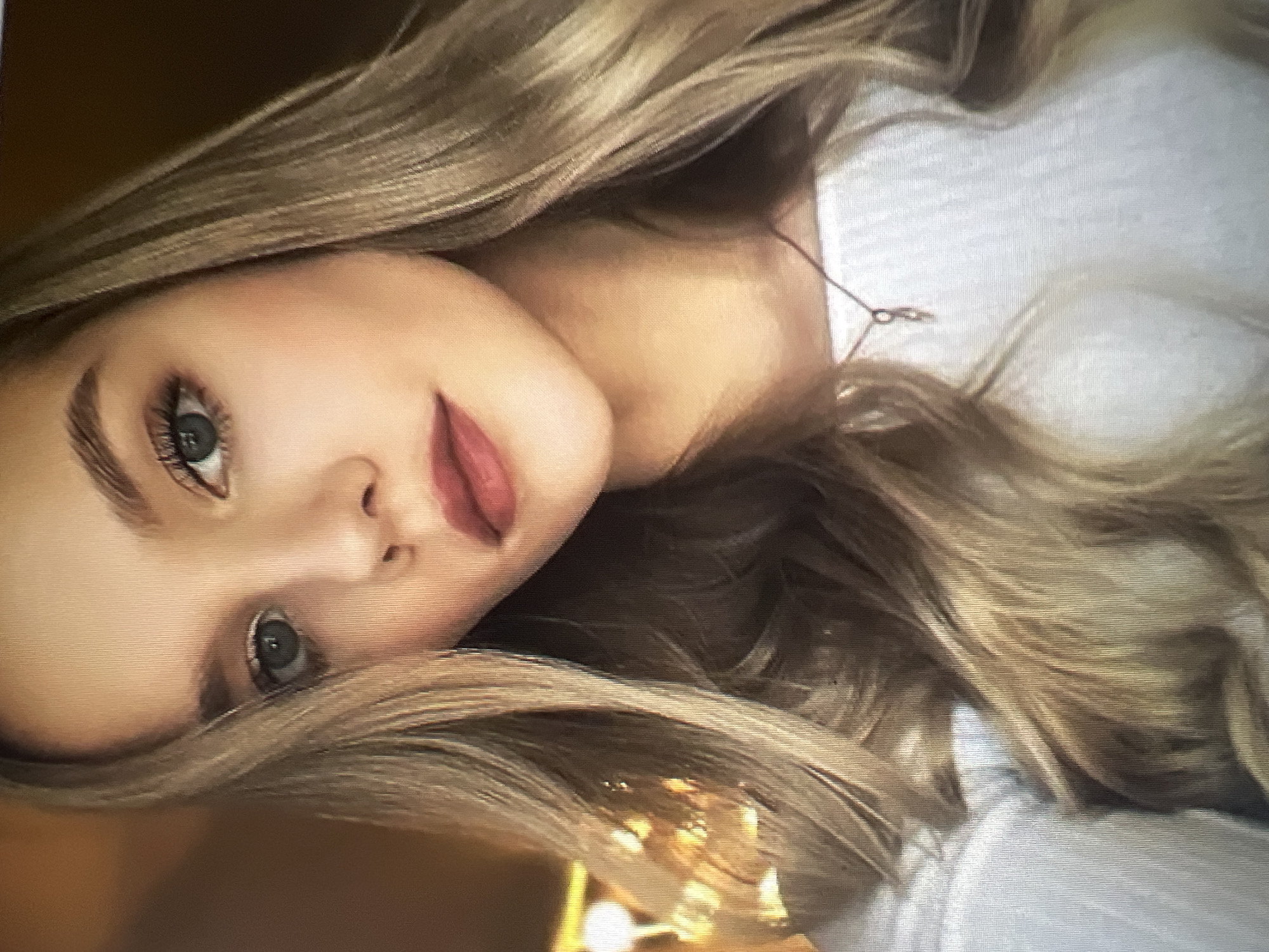 The image shows a close-up, head-and-shoulders portrait of a young woman with blonde hair. She is looking directly at the camera. Her makeup includes mascara, eyeshadow, and lipstick. She is wearing a white top and a delicate necklace with a small pendant. The background is out of focus, with warm, diffused lighting suggesting an indoor setting. There is no discernible information in the image that could indicate a location within the city administration of Qarağandı, Kazakhstan.