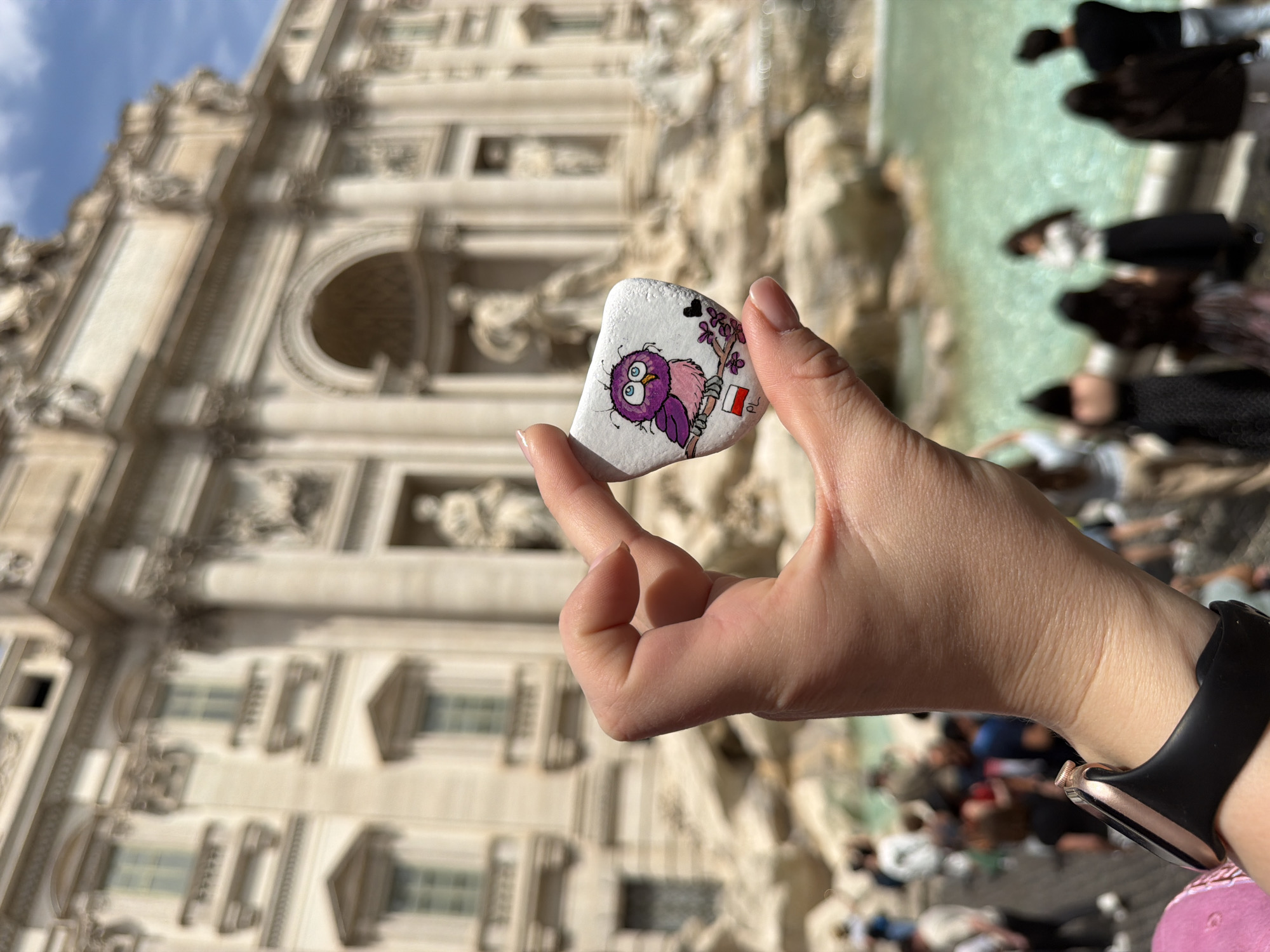 A close-up photograph captures a person's hand holding a decorated stone, set against the iconic backdrop of the Trevi Fountain in Rome, Italy. The irregularly shaped white stone features a colorful, hand-painted design of a whimsical purple and pink owl with large blue eyes, perched on a branch adorned with small purple flowers. A tiny black heart floats above the owl, and a miniature Polish flag (white over red) is tied to the branch.
The hand, with neatly manicured nails, wears a black smartwatch with a rose gold casing on the wrist. In the blurred background, the grand, ornate Baroque facade of the Trevi Fountain is prominently visible, characterized by its intricate carvings, columns, and arched structures made of light-colored stone. Above the fountain, patches of blue sky with white clouds are seen.
Below the architectural marvel, the greenish water of the fountain's basin is visible, with several indistinct figures of people gathered around, suggesting a bustling tourist location. The scene is illuminated by bright, natural daylight, indicating a sunny day, likely during the mid-morning or afternoon. The image offers a personal perspective of a traveler showcasing a unique souvenir or kindness rock at a famous historical site.