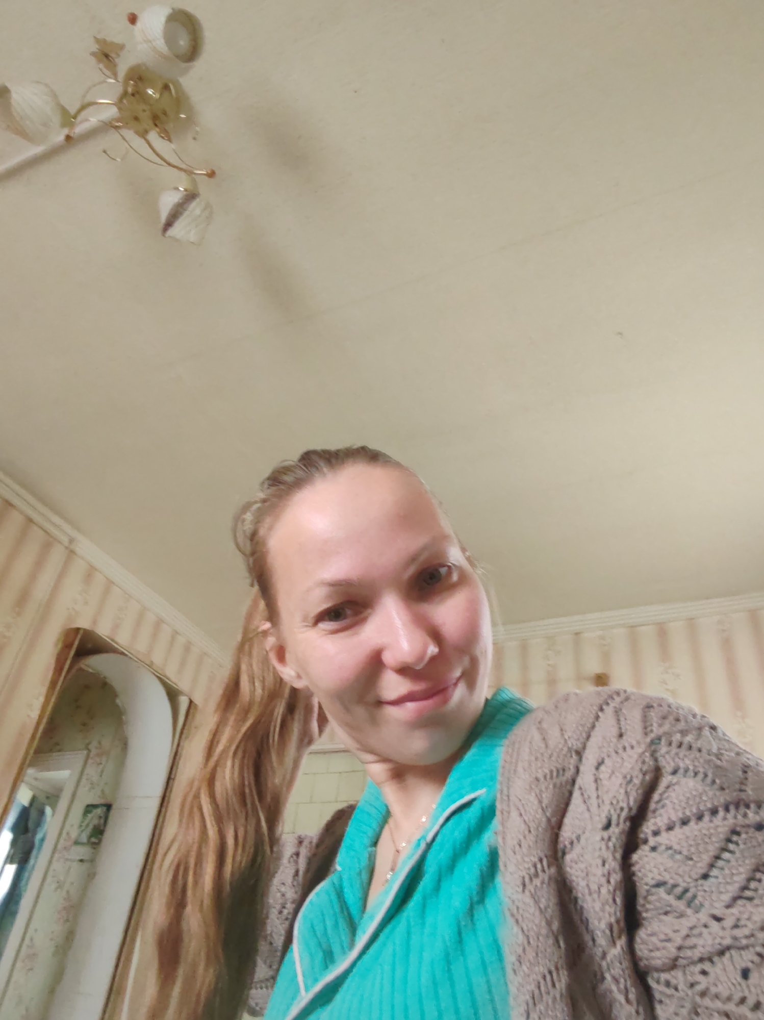 The image displays a selfie of a woman in an indoor setting, likely a residence in Чухлэм, Russia. She has light skin, with light brown hair pulled back into a ponytail, and is captured with a gentle, soft smile, looking towards the camera. She is dressed in a teal-colored top with white trim, layered with a light brown or beige knitted cardigan featuring an openwork pattern. A delicate chain necklace is visible around her neck.
The background shows details of the room's interior. The ceiling is a light cream color with some minor imperfections. A multi-armed chandelier with metallic accents and three light-colored shades (two facing upwards, one downwards) is mounted on the ceiling. The walls are covered with striped wallpaper, featuring alternating wider beige/cream stripes and narrower, patterned stripes, topped by a decorative molding near the ceiling line. On the left side of the image, a tall mirror with a light-colored frame reflects a portion of the room, including what appears to be an arched doorway or structural element and another wall with a lighter, possibly floral-patterned wallpaper and a framed object. The overall ambiance suggests a cozy, traditional interior. No text is visible in the image.