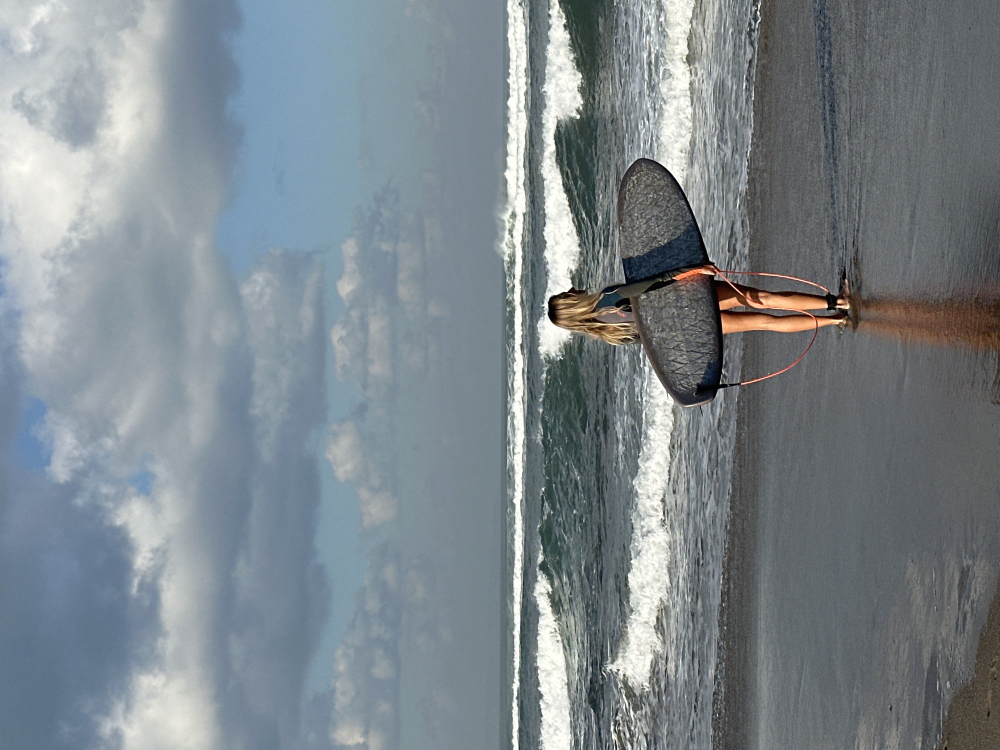 An outdoor, eye-level photograph depicts a lone female surfer standing on a dark, wet sandy beach in North Kuta, Indonesia, observing the ocean. The subject, positioned in the lower-middle frame, has long, blonde hair, wears a dark wetsuit top and dark shorts, and is holding a dark, textured surfboard under her left arm. An orange leash extends from the surfboard to her left ankle. She is barefoot, with her legs reflecting on the damp sand. Her posture indicates she is looking out towards the multiple lines of breaking waves in the dark blue-green ocean. The sky above is partially covered with grey and white clouds, with patches of clear blue sky visible, particularly on the right side. The horizon line is distinct, with a faint, distant landmass or atmospheric haze.