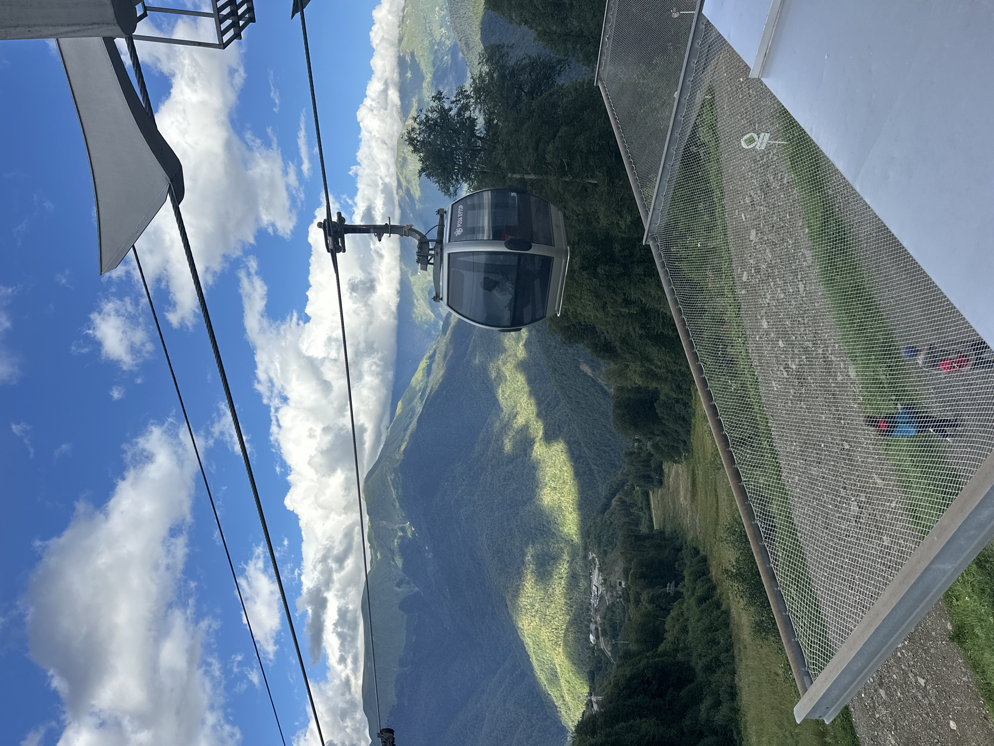 The image captures a scene from a cable car in Estosadok, Russia. The cable car, a modern gondola with tinted windows, is suspended from thick steel cables against a backdrop of lush, green mountains under a partly cloudy sky. Sunlight casts dramatic shadows across the mountain slopes, highlighting a ski slope or trail cutting through the dense forest. In the foreground, a safety net is visible, with two individuals in the lower right corner, appearing to be moving away from the camera. The cable car itself has "РОЗА ХУТОР" branding visible on its side. The overall environment is mountainous and appears to be a resort area, likely during the summer or shoulder season given the green foliage.