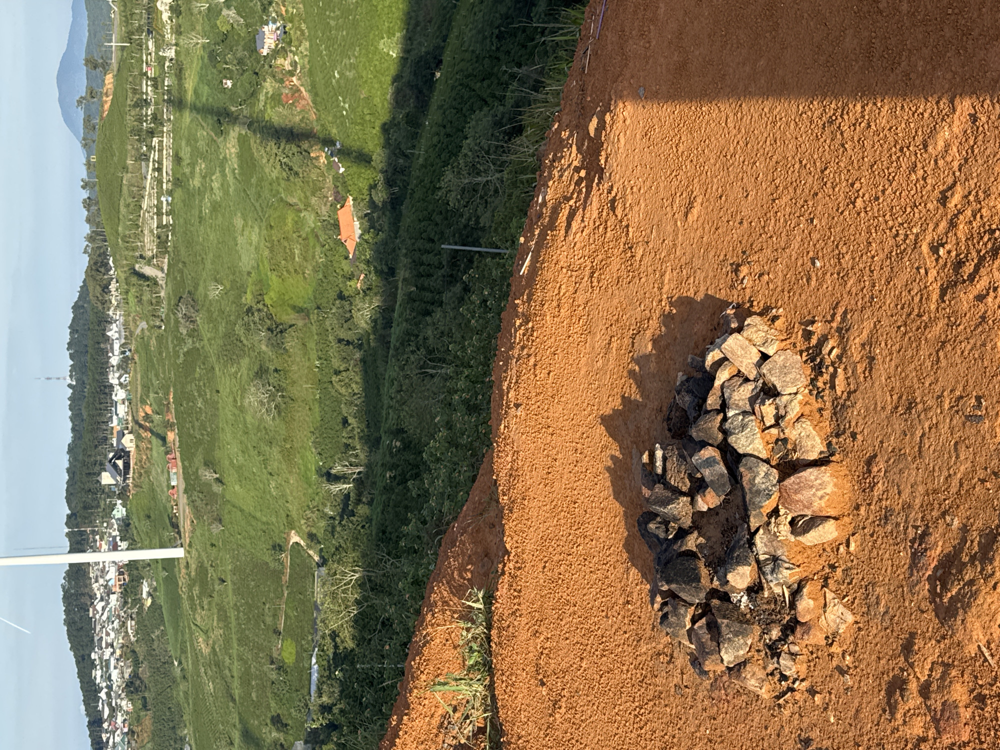 The image depicts a rural landscape in Trường Thọ, Vietnam, characterized by rolling green hills covered in vegetation, likely tea plantations, interspersed with scattered houses and buildings. In the foreground, a circular arrangement of stones sits on a patch of reddish-brown soil, possibly a fire pit. Beyond this, the land slopes upwards, revealing more cultivated hills and a white wind turbine. A town or village is visible on the highest hill in the background, with a dense cluster of houses. The sky is a pale blue, indicating daylight. The scene appears peaceful and natural, with no people or animals present. The overall impression is one of rural tranquility and agricultural productivity.