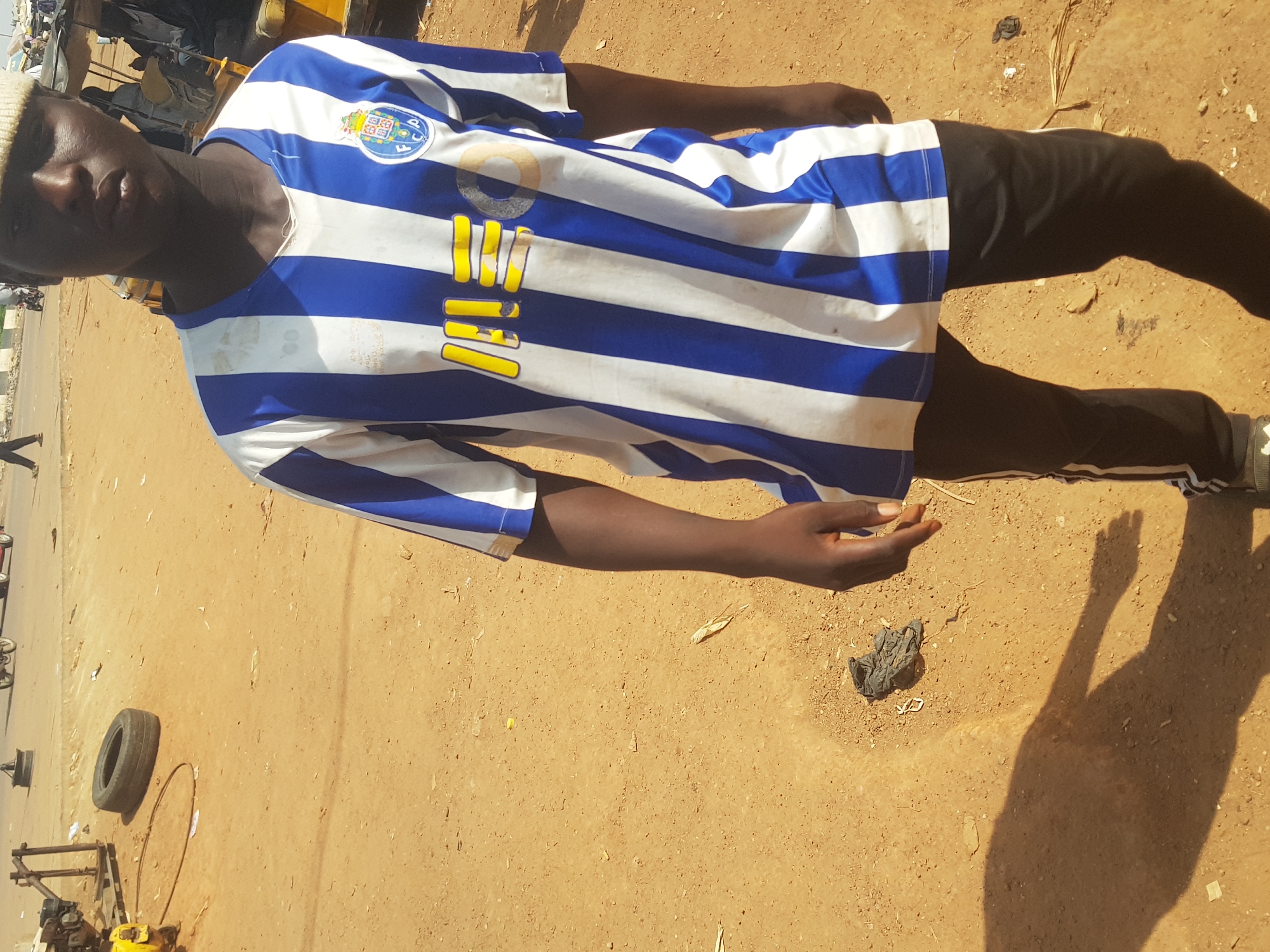 An outdoor, portrait-oriented image captures a dark-skinned individual standing on a dry, sandy, light-brown ground under direct sunlight in Tukur Tukur, Nigeria. The strong, elongated shadows suggest it is either morning or late afternoon.
The subject, likely male, wears a white beanie and a blue and white striped long-sleeved football jersey. On the left chest, a circular crest is visible, identifiable as the emblem of FC Porto, with "FC P" clearly legible within it. Below the collar, several vertical yellow elements, possibly part of a sponsor logo or numbering, are partially obscured and appear worn. The person is also wearing black trousers with white stripes along the side seams. They stand facing slightly towards the camera, with a neutral expression, and their left arm is bent, hand open with the palm facing upwards.
The ground is unpaved and uneven. Scattered objects include a black tire, a metal hoop, pieces of dark plastic or fabric, and a dried plant stem. In the background, an unpaved road stretches into the distance, with vague shapes of vehicles or structures and the legs of another person visible on the far left.