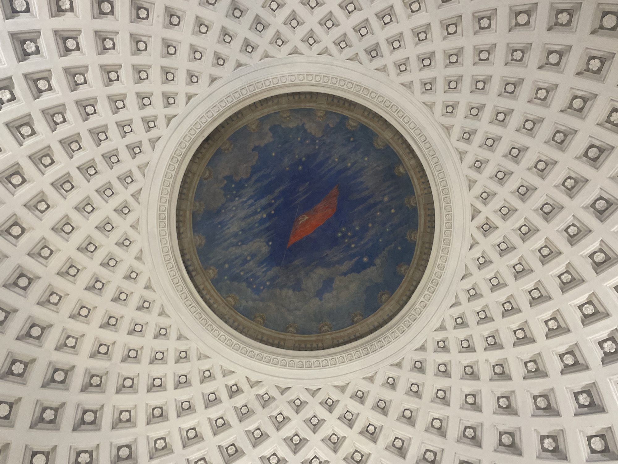 The image shows the ceiling of the mausoleum of Vladimir Lenin in Moscow, Russia. The ceiling has a coffered design with a central circular area containing a mural of a red flag against a blue sky with stars and clouds. The coffers are arranged in concentric circles around the central mural, and each coffer contains a small decorative element, some of which are floral. The image is taken from below, looking directly up at the ceiling.