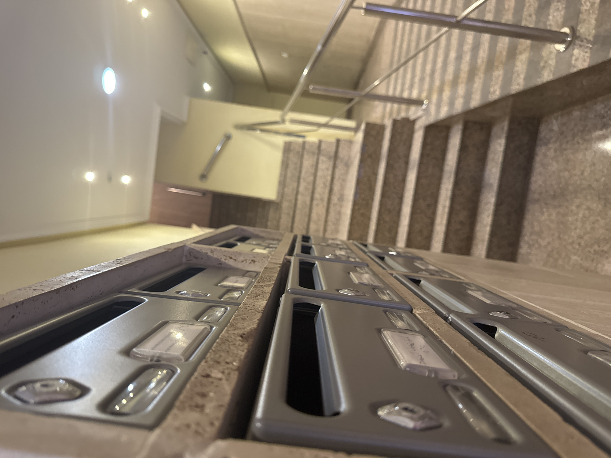 The image depicts an interior hallway or common area with a staircase. In the foreground, rows of grey mailboxes are visible, mounted on a beige speckled surface. Each mailbox has a small window for labels or keys. The mailboxes are arranged in multiple tiers.
The background features a staircase with granite-patterned steps, lined with a silver metal handrail. The walls are painted a pale yellow, and the ceiling has several recessed light fixtures. The overall impression is of a residential building or apartment complex. The image is captured from a low angle, looking up towards the staircase and ceiling.
The prompt requests the location to be the city of Murcia, Spain. However, there is no visual information within the image that definitively places it in Murcia, Spain.