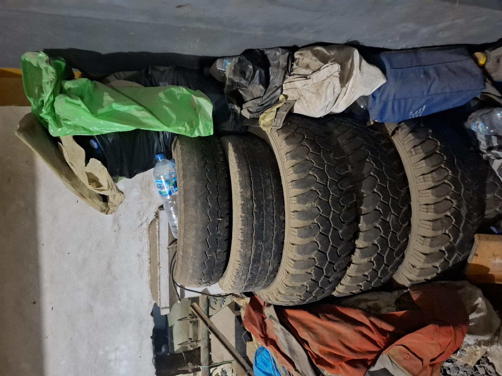 The image depicts a cluttered storage area, likely a garage or shed, located in Ciheulang Tonggoh, Indonesia. In the foreground, a stack of four used tires are visible, covered in dust. To the left of the tires, a pile of assorted items includes a bright orange jacket, some blue fabric, and other unidentifiable materials. Behind the tires, to the right, are several black trash bags, one of which is partially covered by a large green plastic bag. A beige sack and a blue bag are also stacked in this area. A partially filled plastic water bottle rests on top of the uppermost tire. The background consists of a rough, white-washed wall with a damaged section. The overall impression is one of disarray and neglected storage.