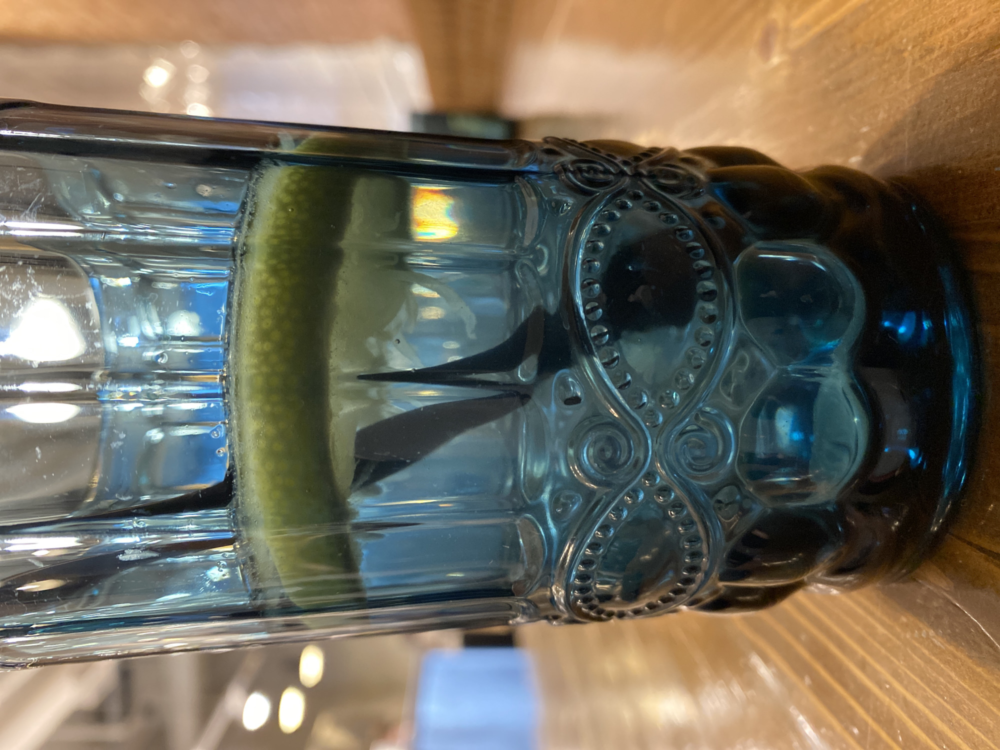 A close-up, sideways shot of a filled blue glass with a lemon slice. A black straw is present inside the glass. The glass sits on a wooden surface. The glass has decorative features. No people are visible. Location is not discernible.