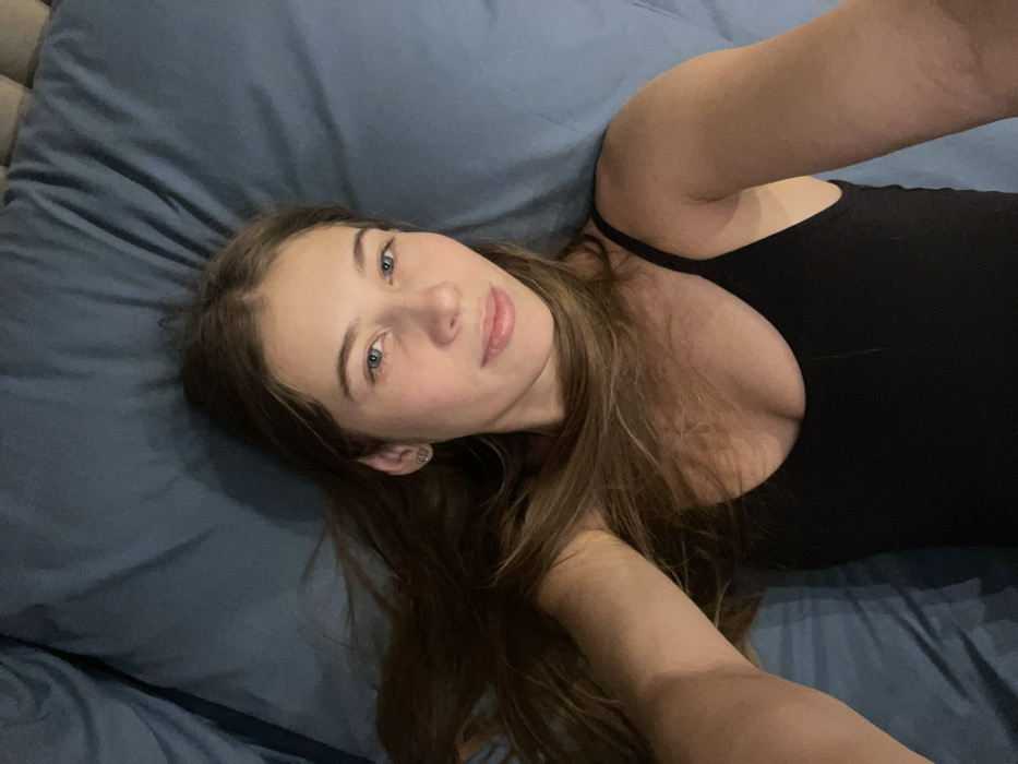 A young woman with long brown hair and blue eyes lies on a blue bed. She is wearing a black tank top. The photo is taken from a high angle, giving a selfie perspective. Her face is turned slightly to the right, and she has a neutral expression. The background is a blue pillow or bed sheet.