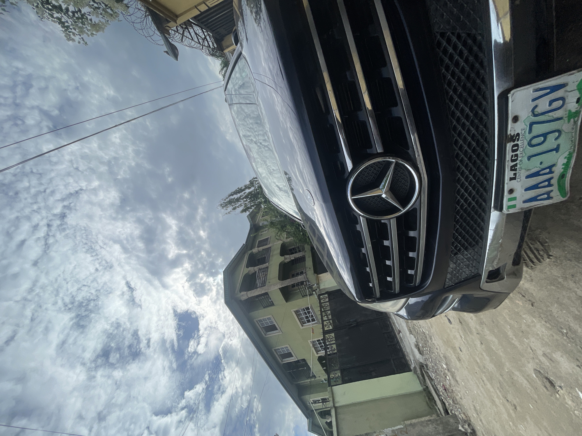 An outdoor daytime scene captures a dark-colored Mercedes-Benz vehicle, likely an SUV or large sedan, in the foreground. The car is positioned at an angle, showcasing its front grille with the prominent Mercedes-Benz star emblem. A Nigerian license plate is partially visible on the front bumper, displaying "LAGOS," "CENTRE OF EXCELLENCE," and the alphanumeric code "AAA-197GY."
In the background, a multi-story building with a light green or pale yellow exterior and numerous windows is visible to the left of the car. The sky above is filled with scattered white and grey clouds, indicating bright daylight. Utility wires traverse the upper part of the image. Along the top edge, barbed wire can be seen, suggesting a security feature on a wall or fence. The ground where the car is situated appears to be a concrete or paved surface. The image depicts a quiet urban or suburban setting, with no specific activity taking place. The license plate confirms the location as Nigeria.