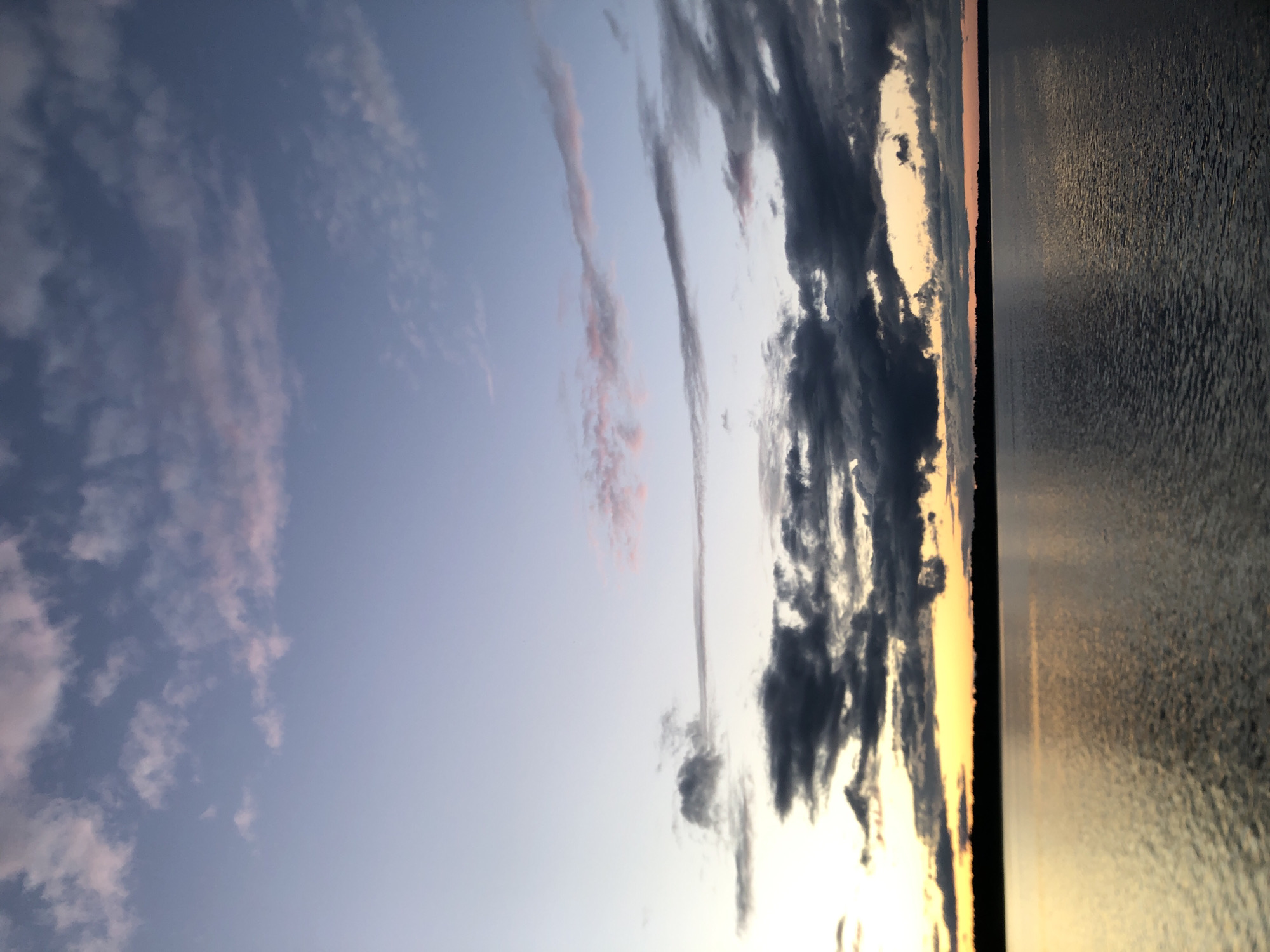 The image depicts a tranquil, natural scene at dusk or dawn, as indicated by the soft, diffused lighting and the hues of the sky. The composition is divided vertically, with the left side featuring a sky filled with wispy clouds tinged with pink and purple against a gradient of pale blue. The right side of the image shows a calm body of water, likely a lake or river, reflecting the warm tones of the sky, creating a shimmering, textured surface. A thin strip of land is visible as a dark silhouette along the horizon, separating the sky from the water. The overall atmosphere is serene and peaceful, evoking a sense of quiet contemplation. No people, objects, or text are visible in the image. The location context of Svirstroy, Russia, suggests a possible setting near a body of water in a rural or natural environment.