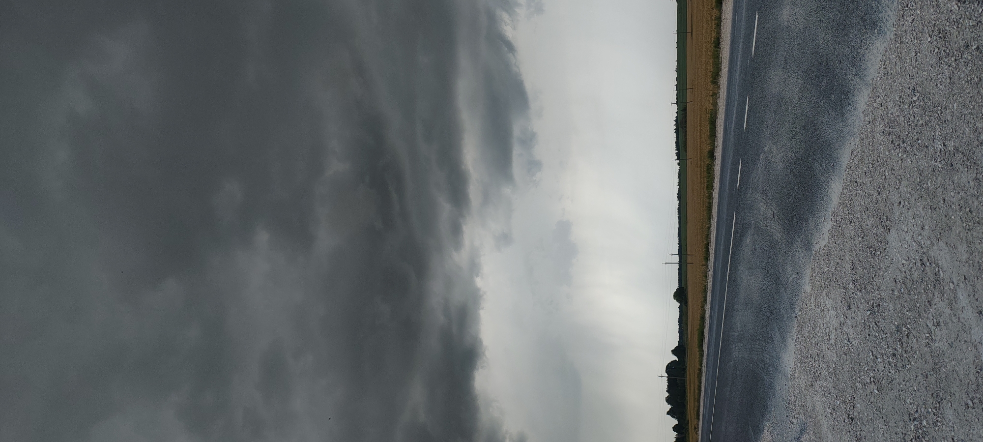 The image depicts an overcast rural landscape with a stormy sky. In the foreground, a wet asphalt road with white lane markings curves to the left, bordered by a gravel shoulder. Beyond the road, a field of dry, yellowish vegetation stretches to the horizon, punctuated by utility poles and a line of trees. A rain shower is visible in the distance, indicated by vertical streaks of precipitation against a bright, hazy sky. The overall atmosphere is somber, suggesting approaching or ongoing bad weather. The location is specified as the city of Новый Изборск, Russia.
