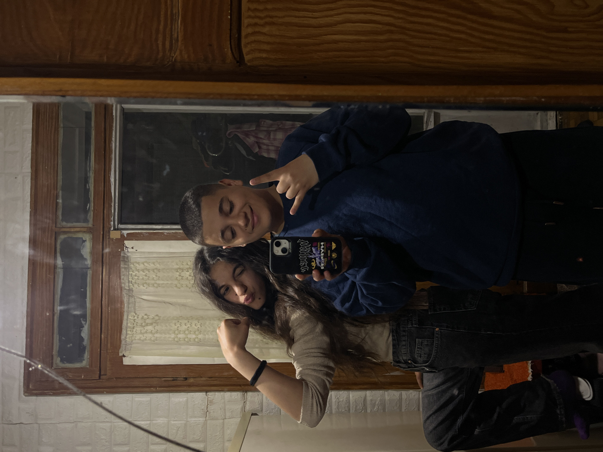 A mirror selfie taken indoors. A boy with short hair and a blue sweater is making a hand gesture. A girl with long hair is flexing her arm. They are holding a phone with a dark case. The background includes a window with a white curtain, wood paneling, and a tiled wall. The location is unspecified within Lozarevo, Bulgaria.