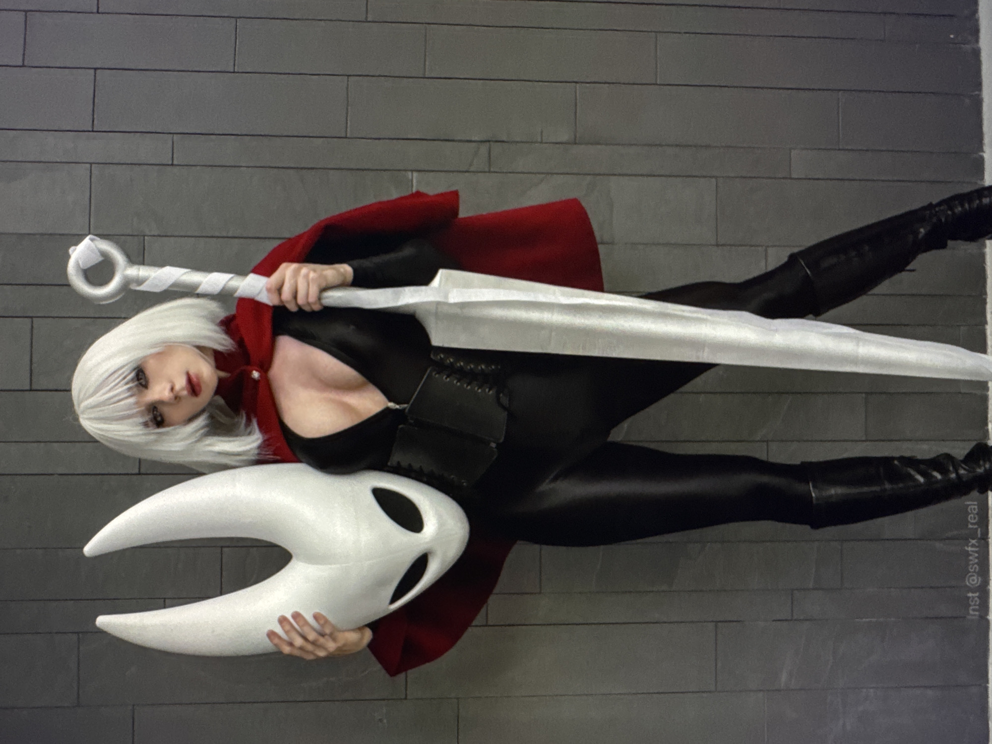 A person stands in front of a gray, tiled wall, holding a large, white, horn-like mask in their left hand and a long, silver sword in their right. The person is wearing a black, skintight outfit with a red cape draped over their shoulders. The mask has two large, black oval eyeholes and two curved horns pointing upwards. The sword is flat and broad, with a circular pommel. The person has short, white hair and is looking towards the camera. A watermark reading "Inst @swfx_real" is visible at the bottom of the image. The scene appears to be indoors, possibly a studio or a home setting.