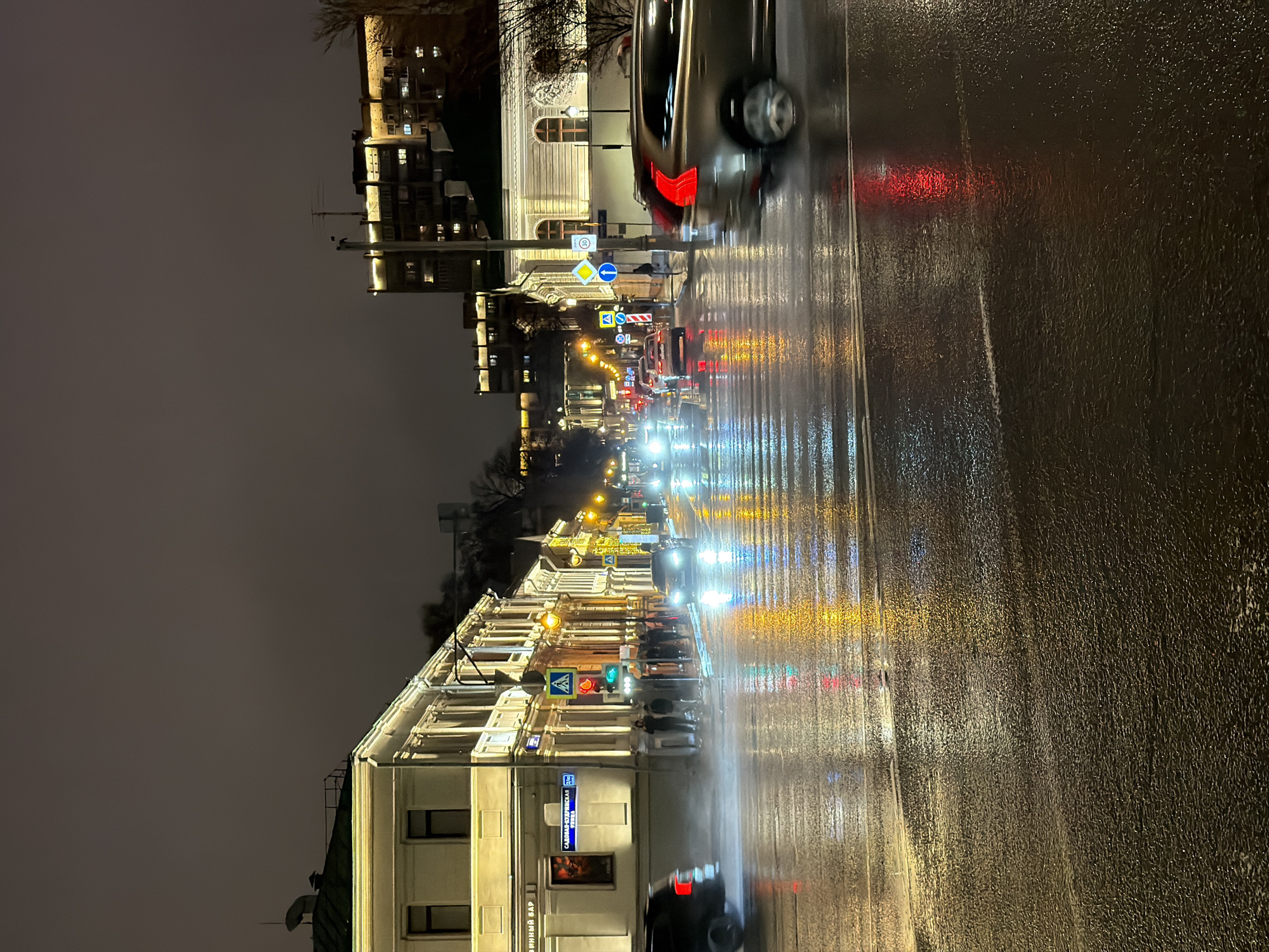 The image depicts a street scene in Moscow, Russia at night during a rainstorm. The street is wet, with reflections of the streetlights and vehicle lights visible on the asphalt. Numerous cars with their headlights and taillights illuminated are present. Along the street, buildings with lit windows stand on either side. There are several street signs and traffic lights visible. The buildings are predominantly white with architectural details. The sky is dark and overcast.