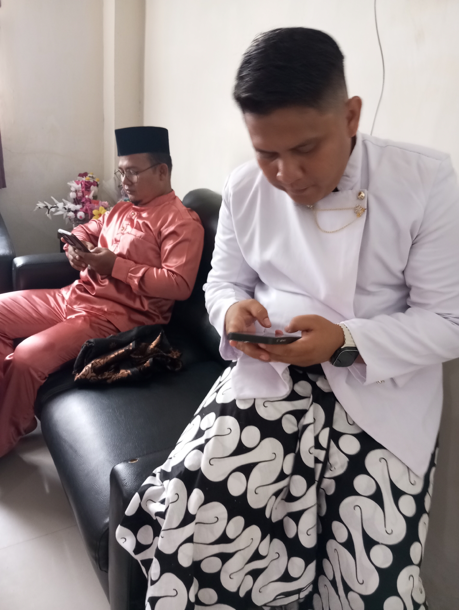 Two men, likely of Indonesian nationality, are seated on a black leather couch in what appears to be a domestic indoor setting, possibly a home in Pematang Reba, Indonesia. The man on the left, wearing a peach-colored traditional Islamic outfit and a black peci (skullcap), is looking down at his mobile phone. He has glasses on and is leaning back on the couch. A folded dark fabric with gold patterns rests on the couch between him and the other man. The man on the right is dressed in a white traditional tunic over a black and white patterned sarong. He is also looking down and interacting with his mobile phone. A gold chain is visible on his chest, attached to his tunic. The background consists of a plain wall and a corner with some decorative artificial flowers. The lighting suggests an indoor environment, possibly with ambient daylight or artificial lighting.