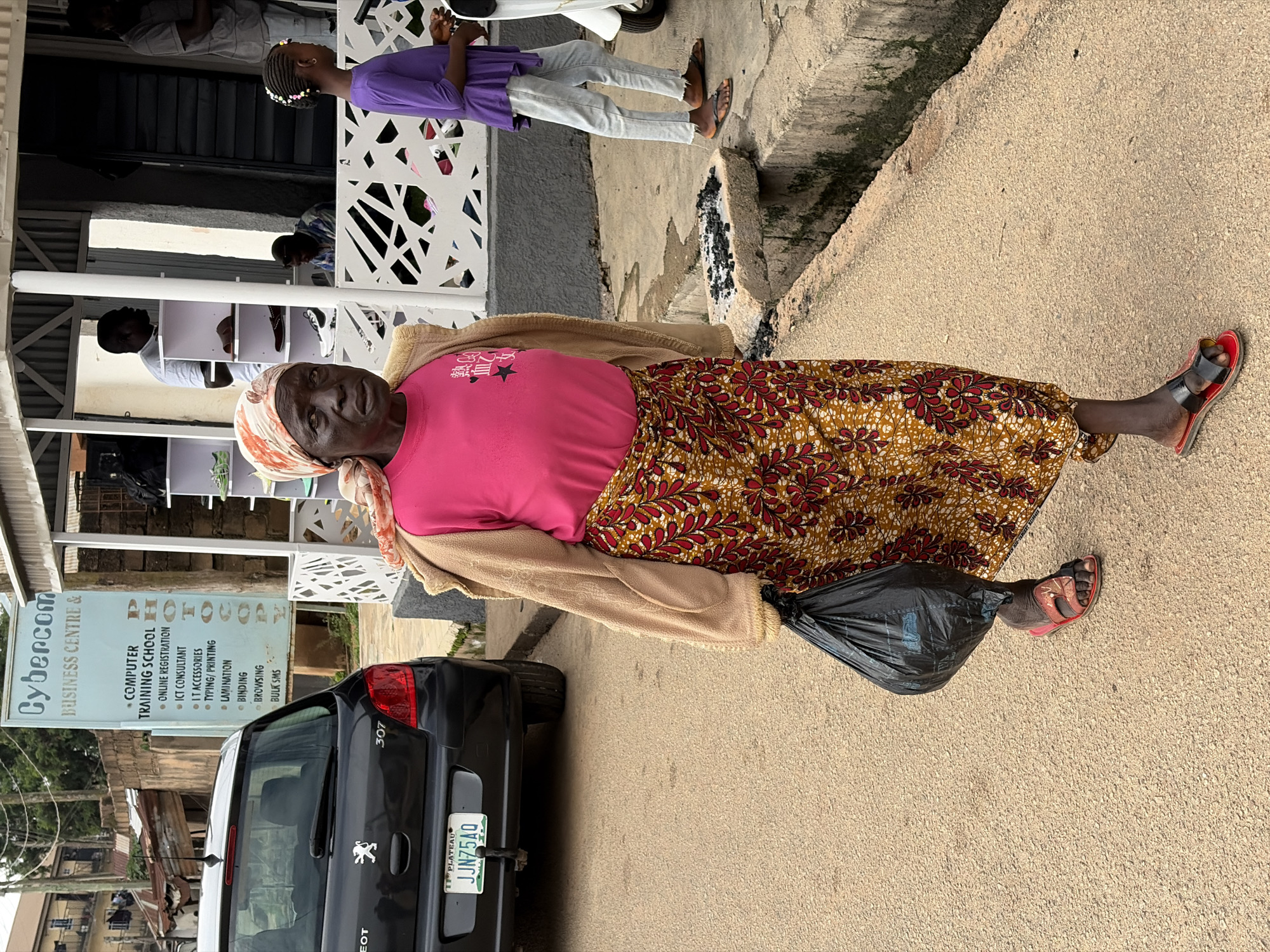 A full-body shot captures an elderly African woman walking down a paved street in Jos, Nigeria. She wears a bright pink t-shirt, a tan cardigan, and a vibrant patterned skirt. Her head is covered with a light orange and white patterned headscarf. In her left hand, she carries a black plastic bag. She is wearing red sandals. To her left, a black Peugeot 307 car is parked on the street. Behind the woman, a sign for "Cybercom Business Centre & Computer Training School" is visible. The area appears to be a commercial district with a building and shops in the background. A young girl in a purple dress and light blue pants walks past the woman in the background. The ground is a textured pavement, and a concrete gutter runs along the right side of the street.