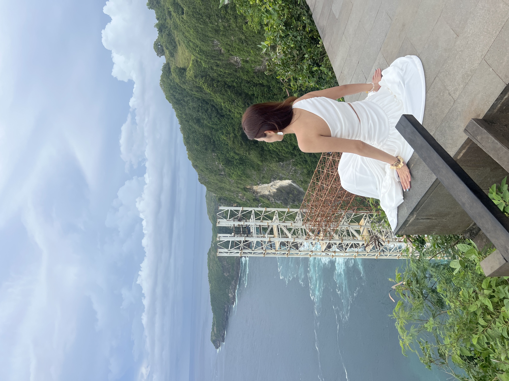 This image captures a scenic view from a high vantage point in Nusa Penida, Indonesia. The central subject is a woman with brown hair, dressed in a white, sleeveless dress with a bare midriff. She is kneeling on a light grey, tiled stone platform, looking out towards the vast ocean and dramatic coastline. She wears a white earring and gold-toned bracelets on her wrists.
The background features towering, verdant green cliffs that drop sharply into the blue-green sea. White foamy waves can be seen crashing against the shoreline. Across the water, a large, complex metal truss structure, possibly an observation platform or part of a construction project, spans the bay. The sky above is largely overcast with soft white and grey clouds, indicating a cloudy but bright daytime. In the immediate foreground on the right, a dark grey railing or ledge is partially visible. The scene conveys a sense of awe and appreciation for the expansive natural beauty. No visible text is present in the image.